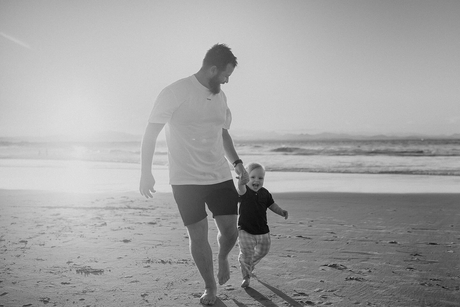 Black and white photo of dad walking with his toddler son along the water's edge