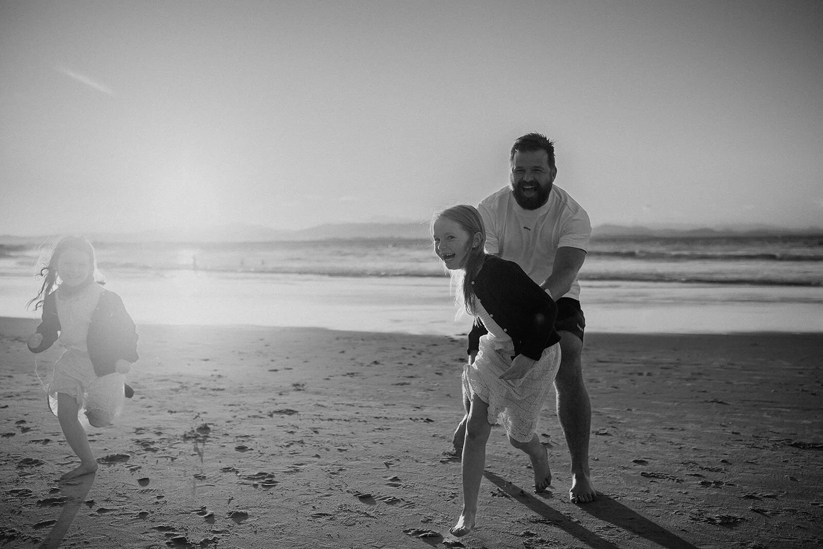 Black and white photo of dad chasing his daughters along the beach