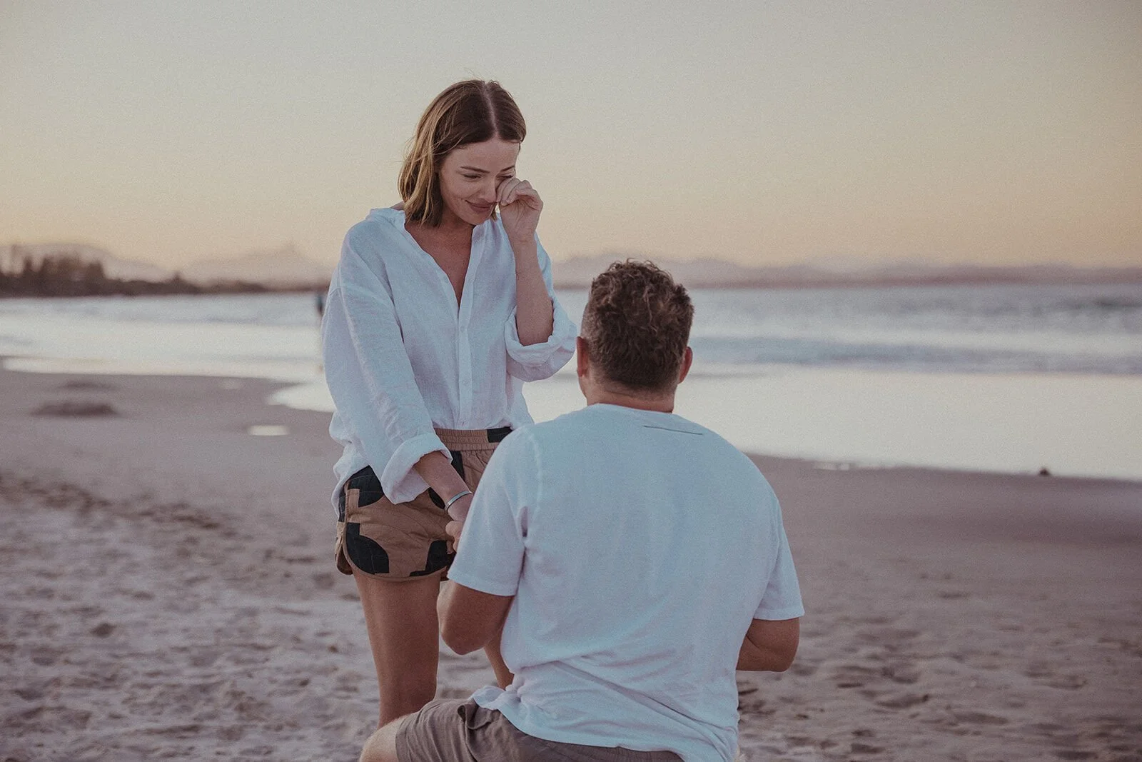 Woman wiping away a happy tear as her partner kneels in front of her, Jamie and Jessica's beach marriage proposal at Belongil Beach, Byron Bay