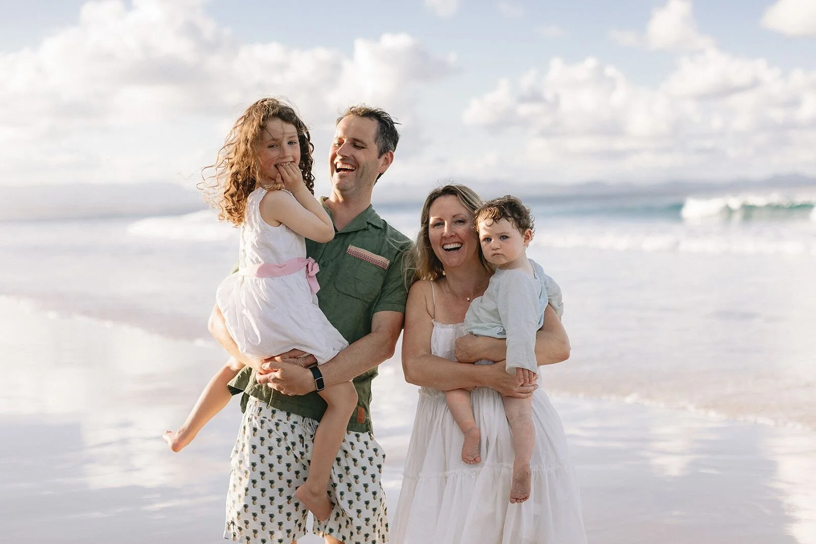 Family of four laughing together at the water's edge, dad lifting his daughter, ocean waves behind