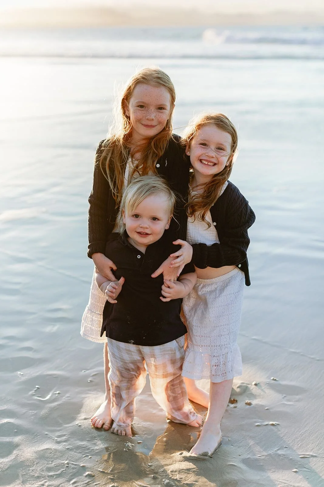 Two sisters hugging their toddler brother between them as they stand in the shallows