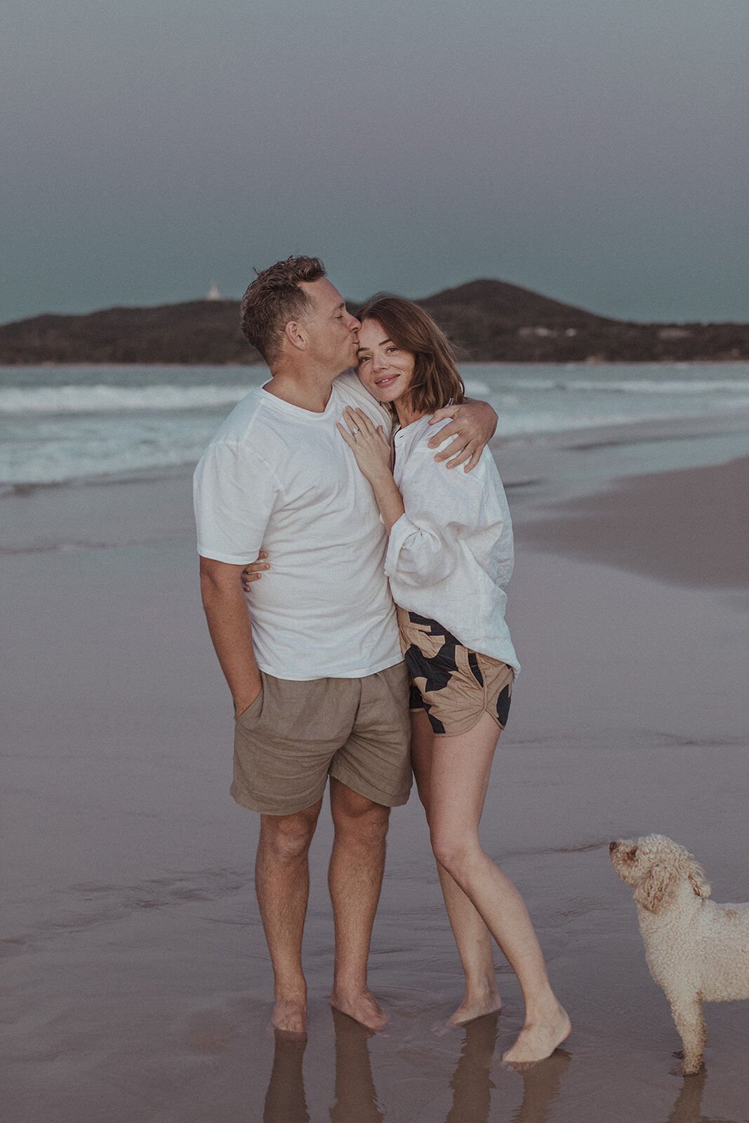 Couple embracing with the Cape Byron headland behind, Jamie and Jessica's beach marriage proposal at Belongil Beach, Byron Bay