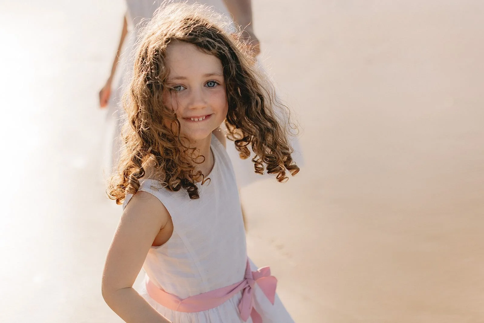 Portrait of a curly-haired girl in a white dress with a pink sash, glowing in golden light