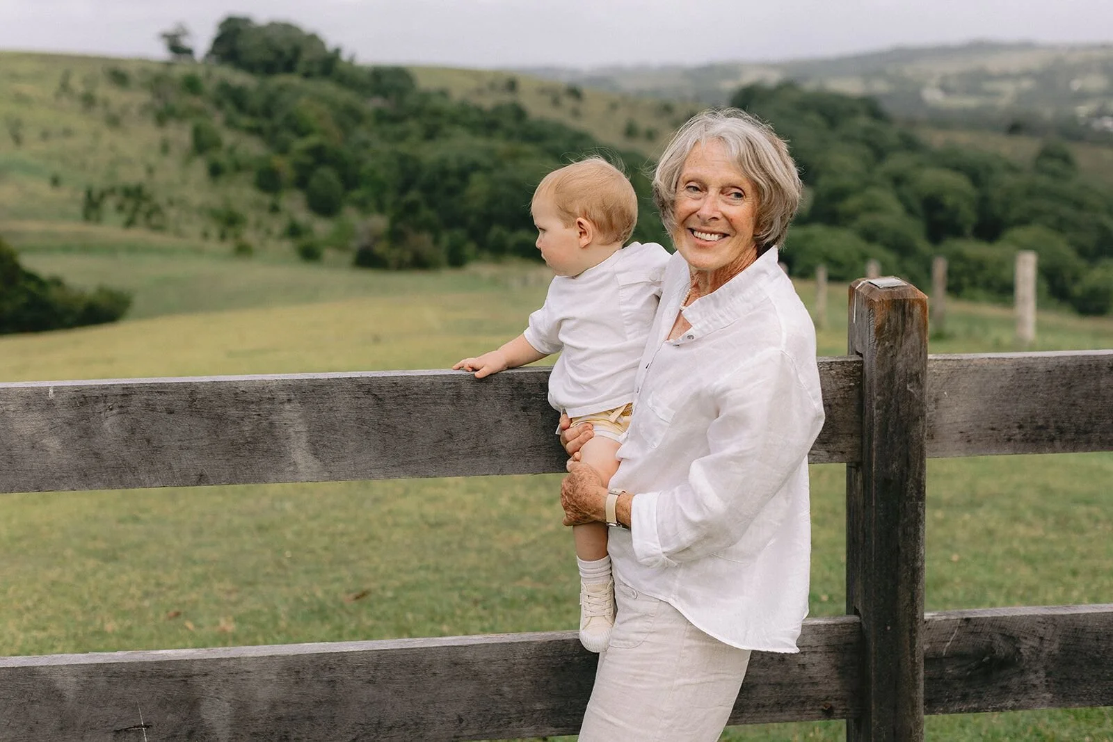 Grandmother holding her baby grandson against a timber fence with the hills behind