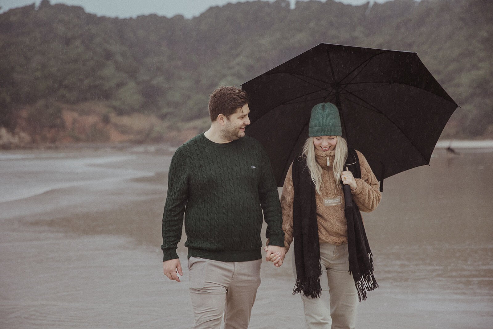 Couple walking hand in hand under the umbrella with the headland behind, Raphael and Vany's rainy day marriage proposal at Broken Head Beach, Byron Bay