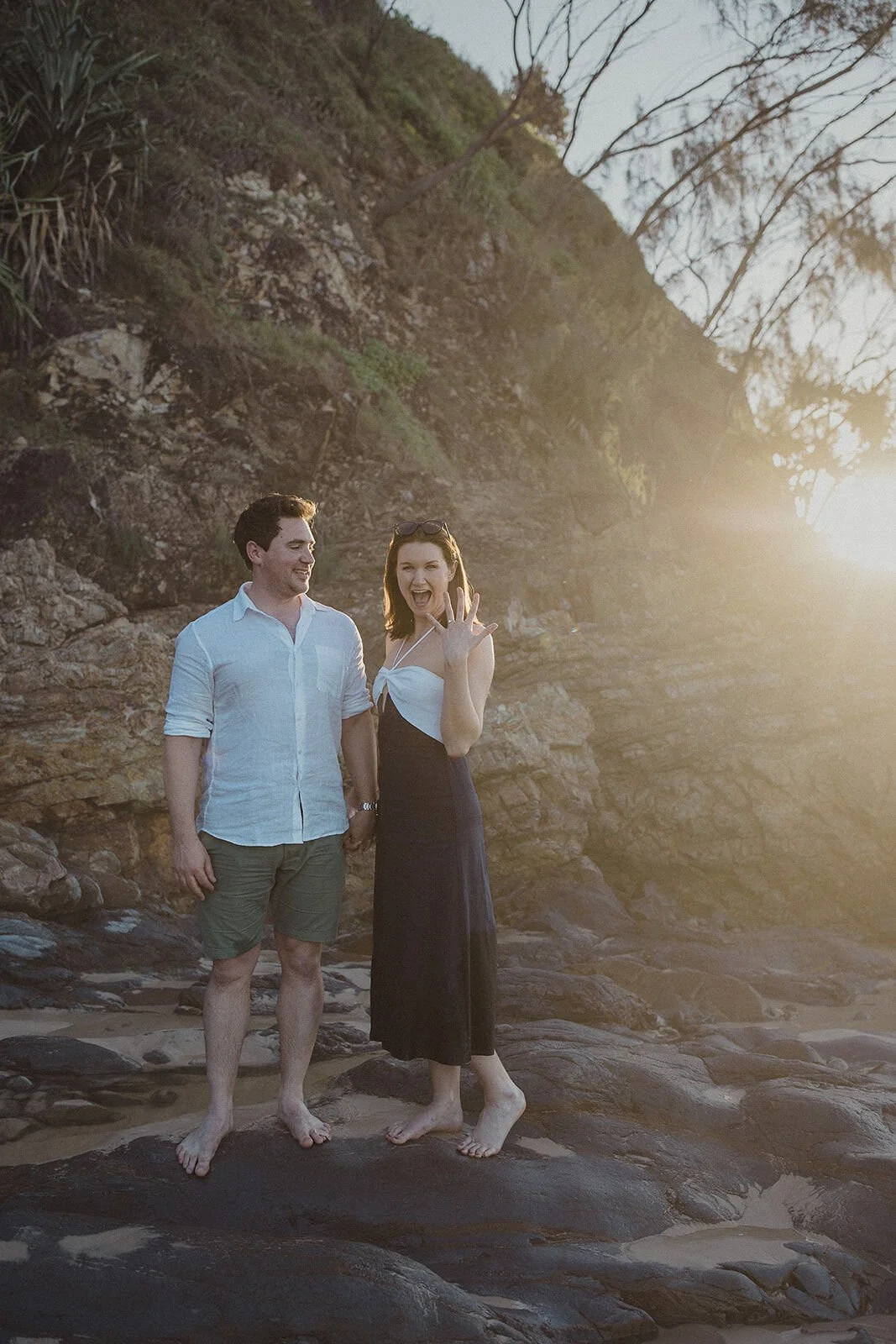 Couple standing on the rocks, the woman showing her ring, John and Emma's sunset marriage proposal at Wategos Beach, Byron Bay