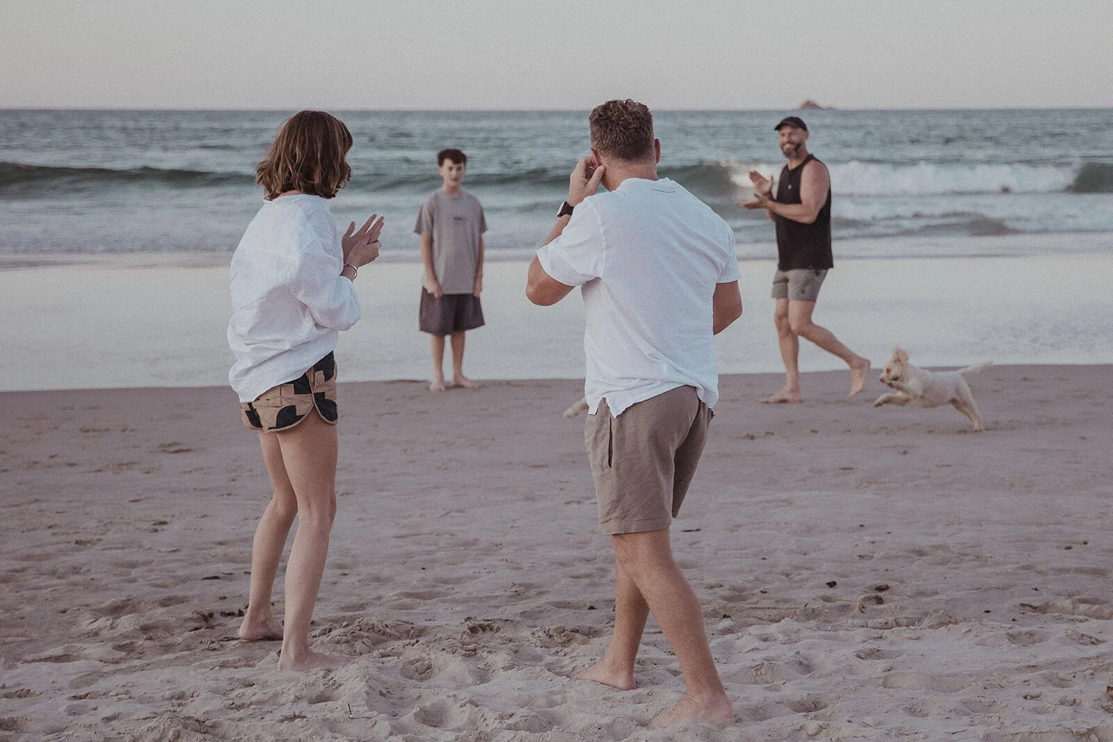 Couple talking while family and friends celebrate in the background, Jamie and Jessica's beach marriage proposal at Belongil Beach, Byron Bay