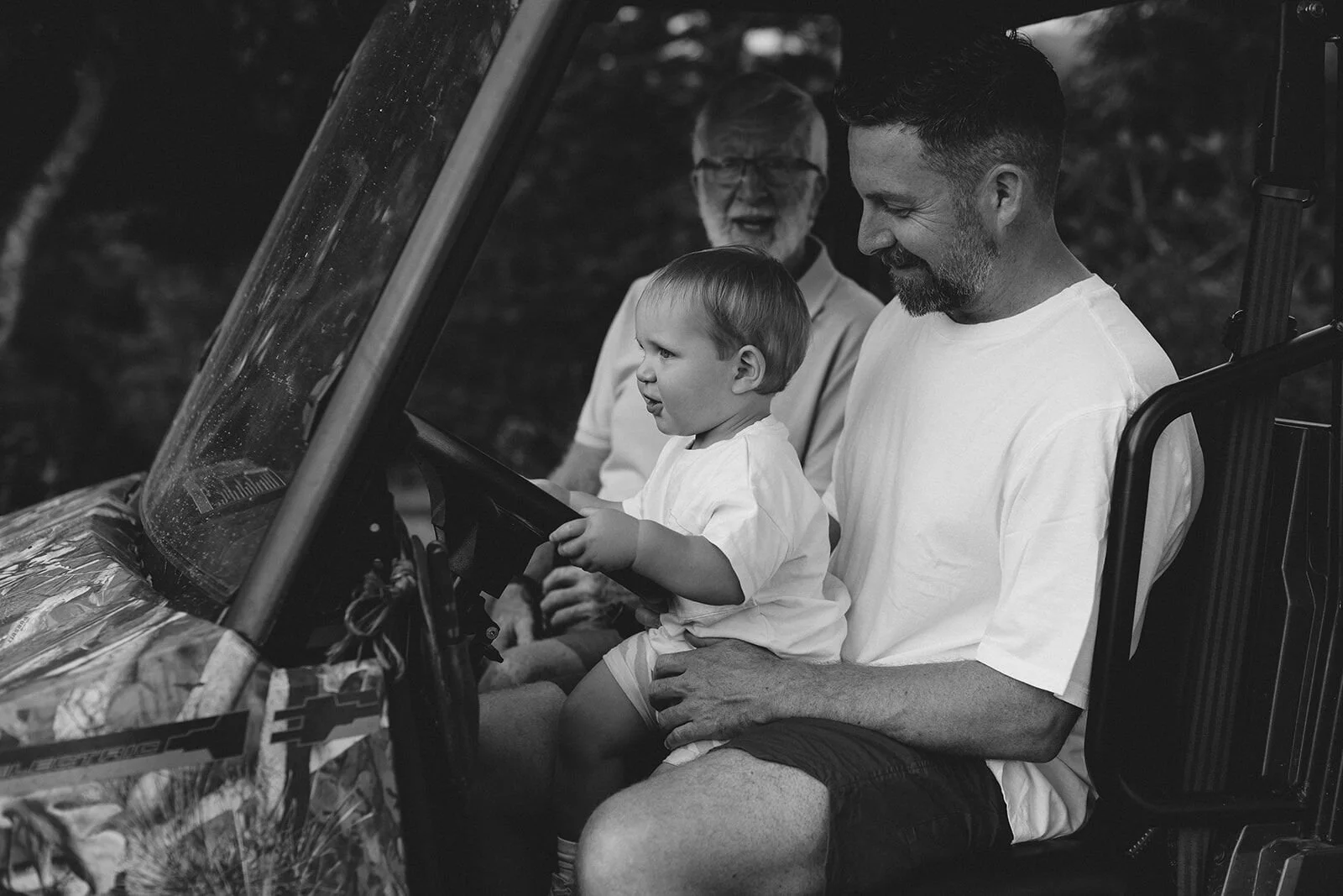 Black and white photo of dad with his toddler son at the wheel of the farm buggy, grandfather behind