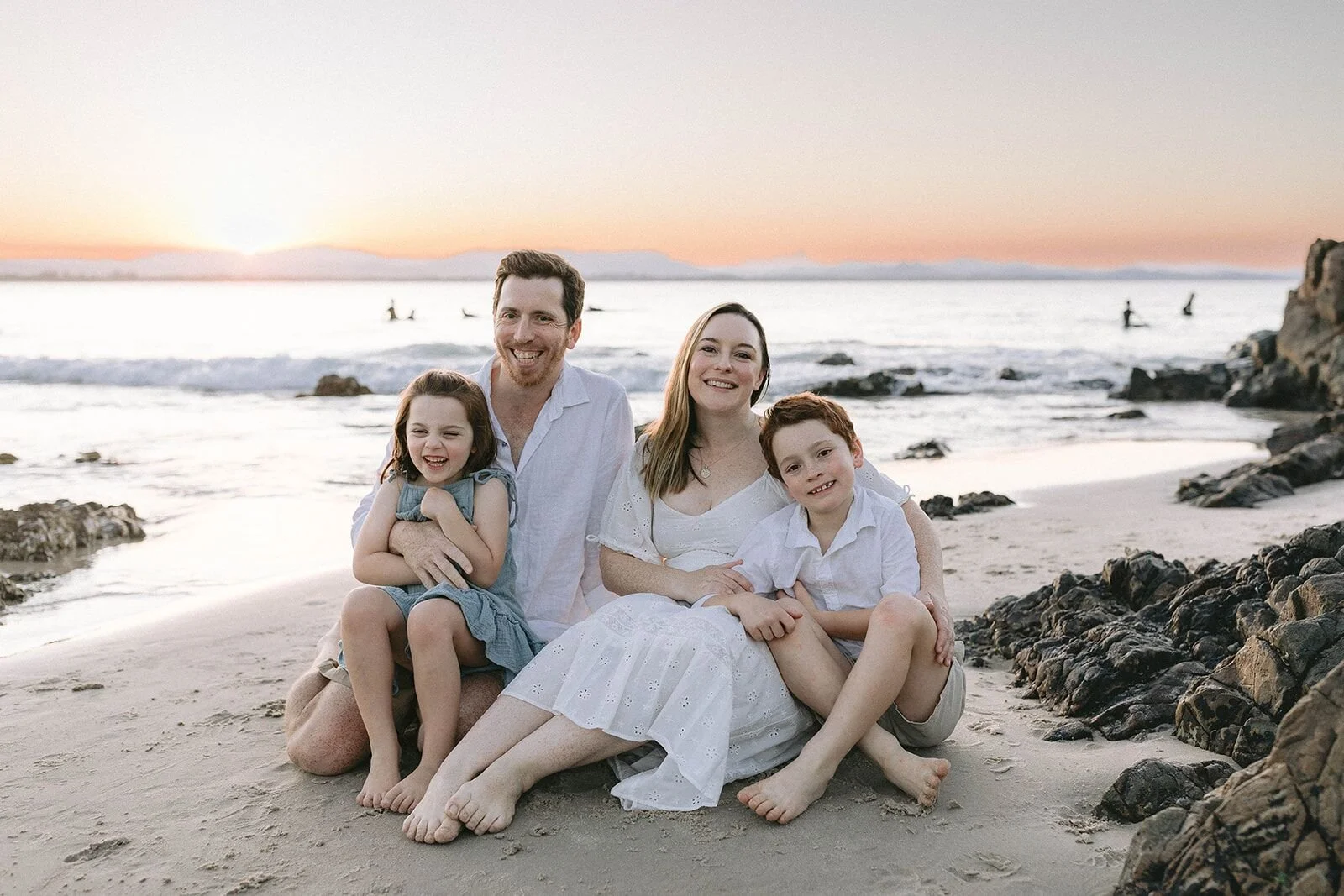 Family of four sitting together on the sand near the rocks as the sun sets