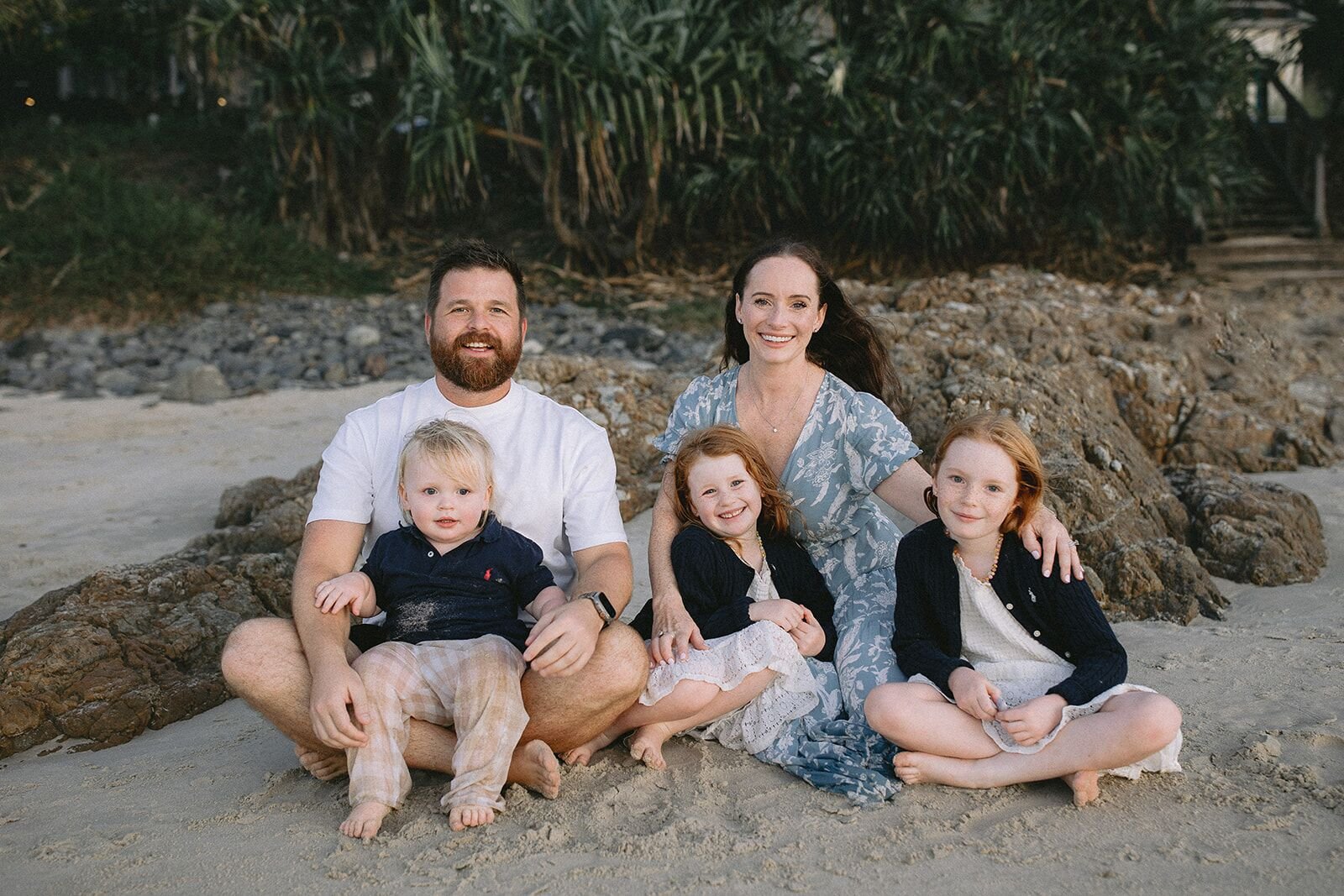 Family of five sitting together on the sand in front of the rocks at Wategos Beach