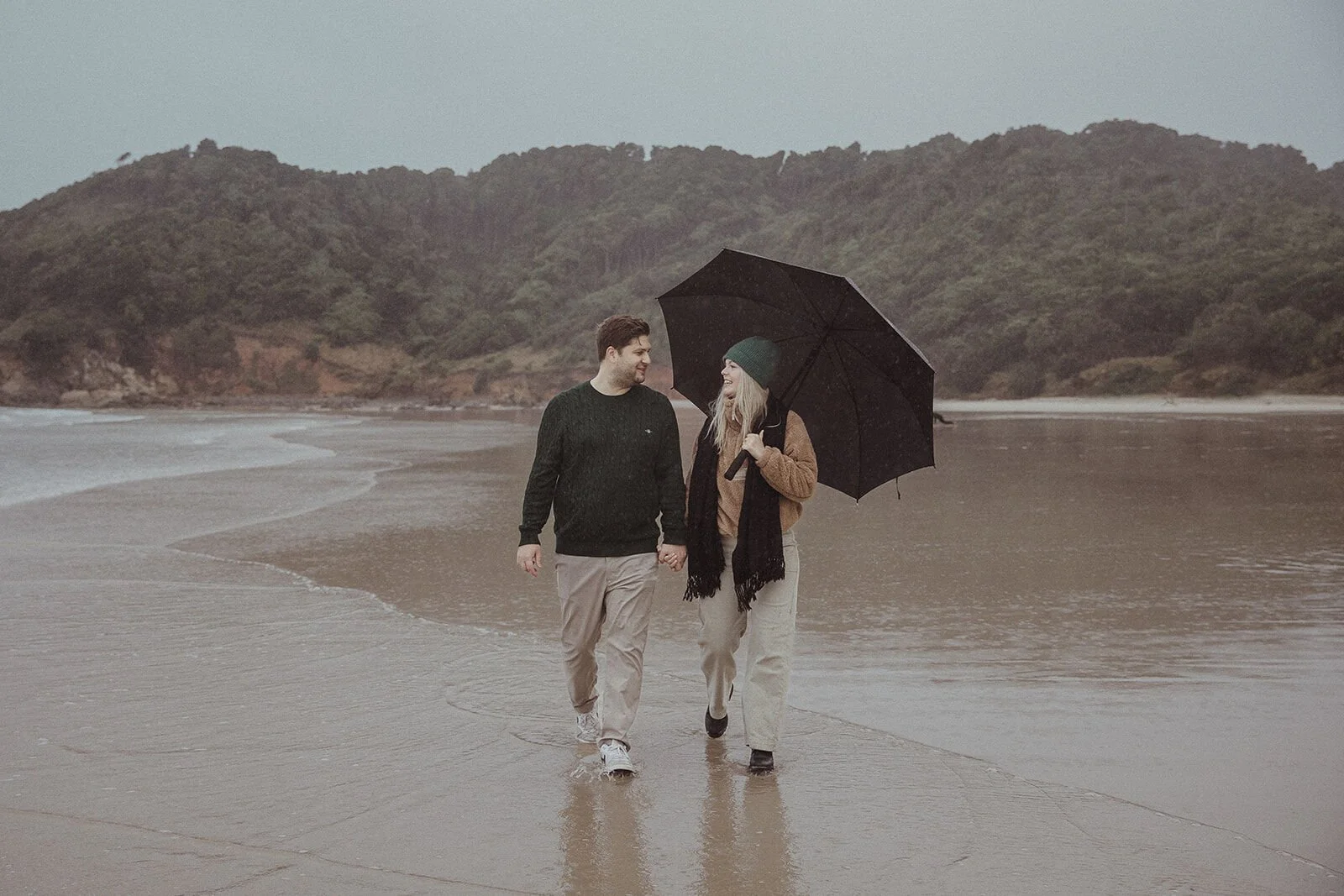 Couple walking along the wet shoreline under an umbrella, Raphael and Vany's rainy day marriage proposal at Broken Head Beach, Byron Bay