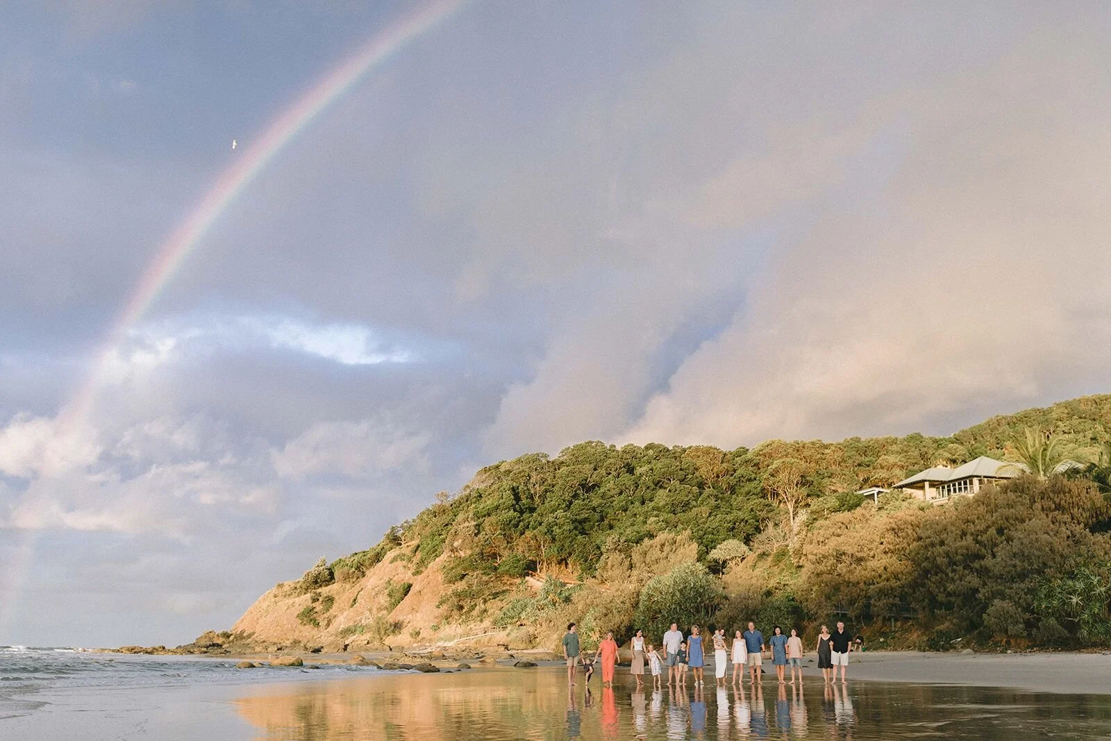 Wide view of a family on the shore under a rainbow, with the green headland of Wategos behind them