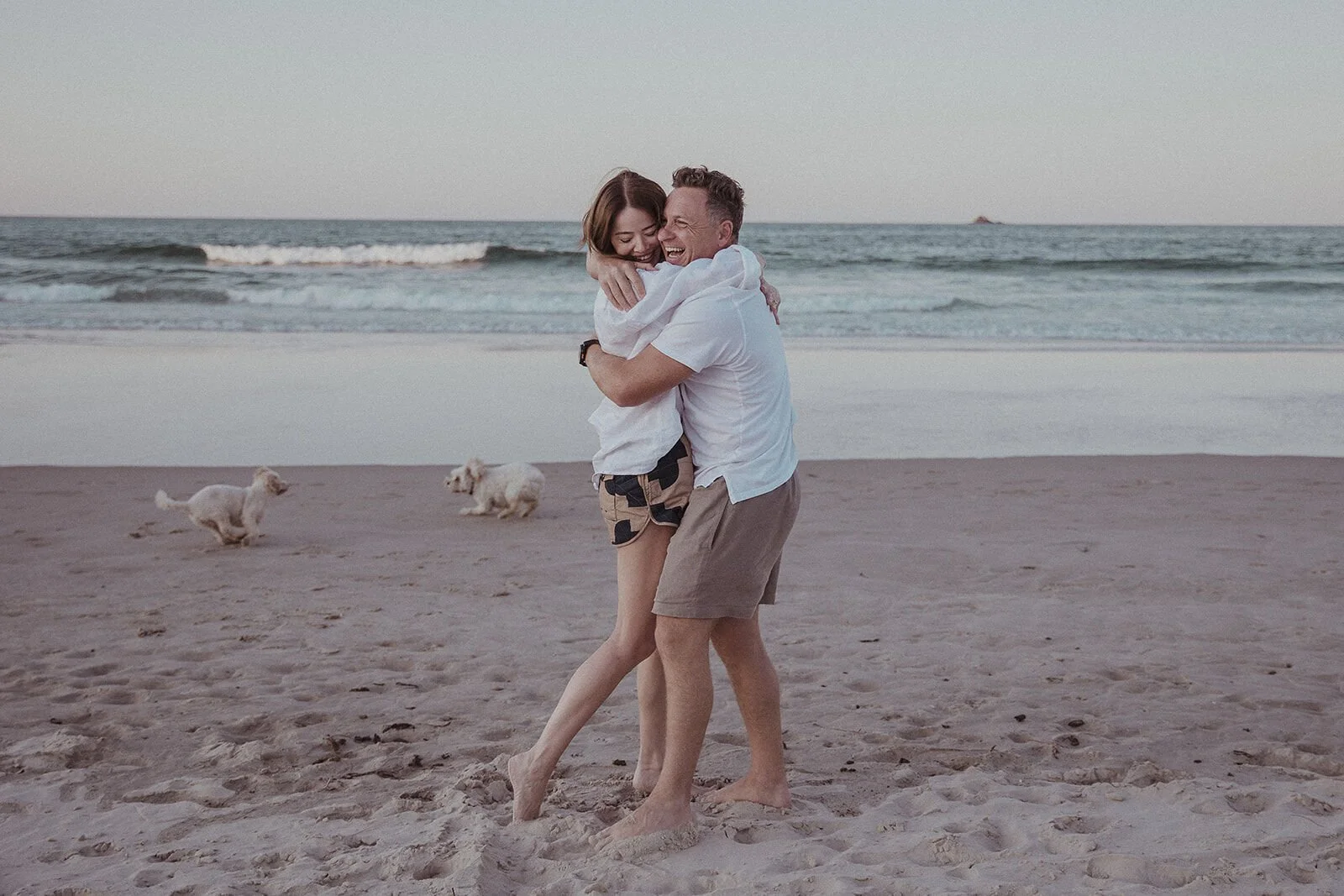 Couple hugging and swinging around on the shore while the dogs play, Jamie and Jessica's beach marriage proposal at Belongil Beach, Byron Bay