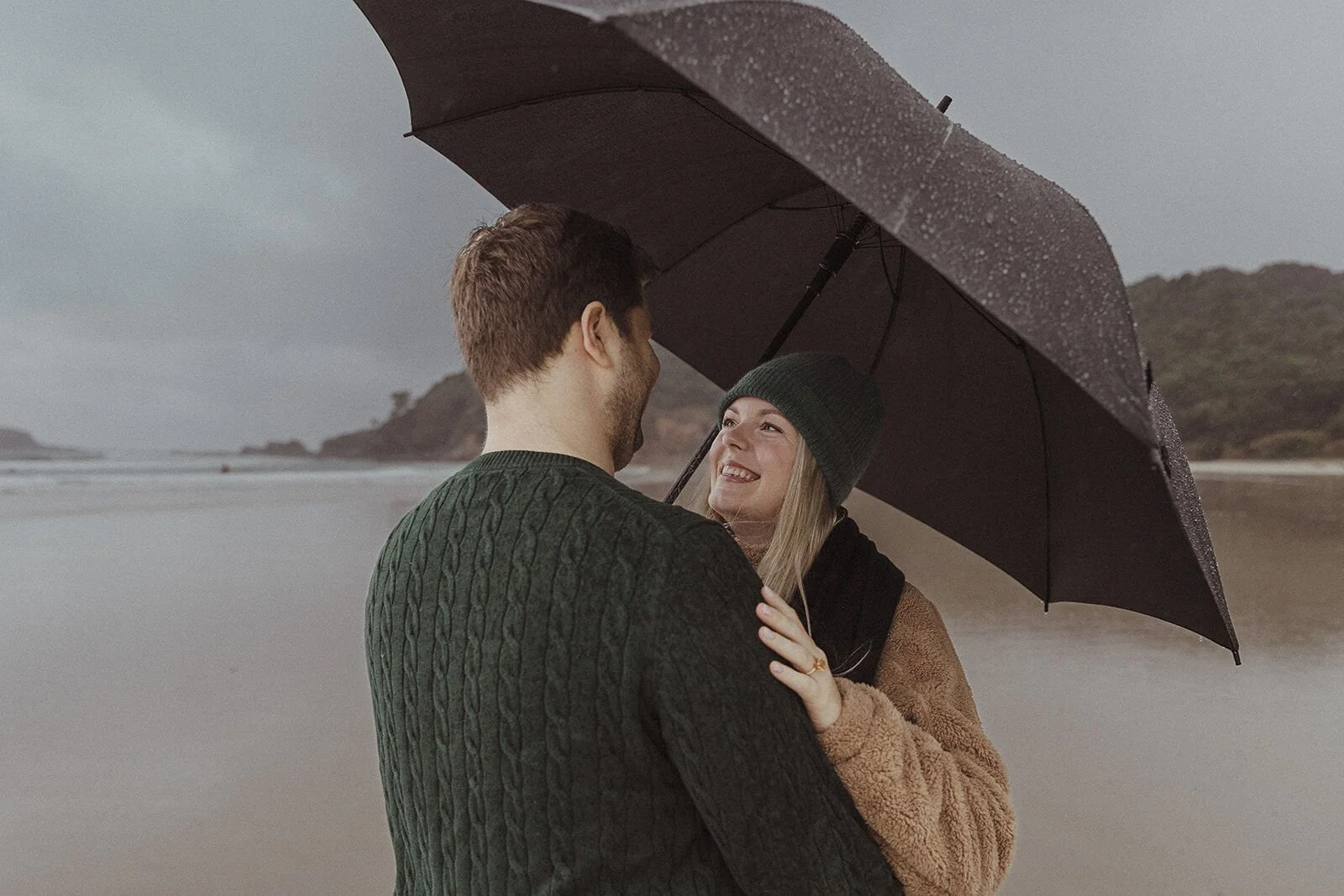 Woman in a green beanie smiling up at her fiancé under the umbrella, Raphael and Vany's rainy day marriage proposal at Broken Head Beach, Byron Bay