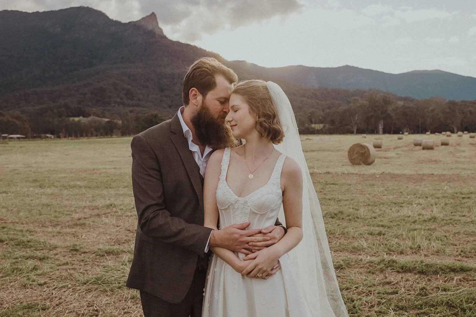 Couple embracing in the hay field with the mountain behind, Sam and Tom's elopement at Highfield Farm, Northern Rivers