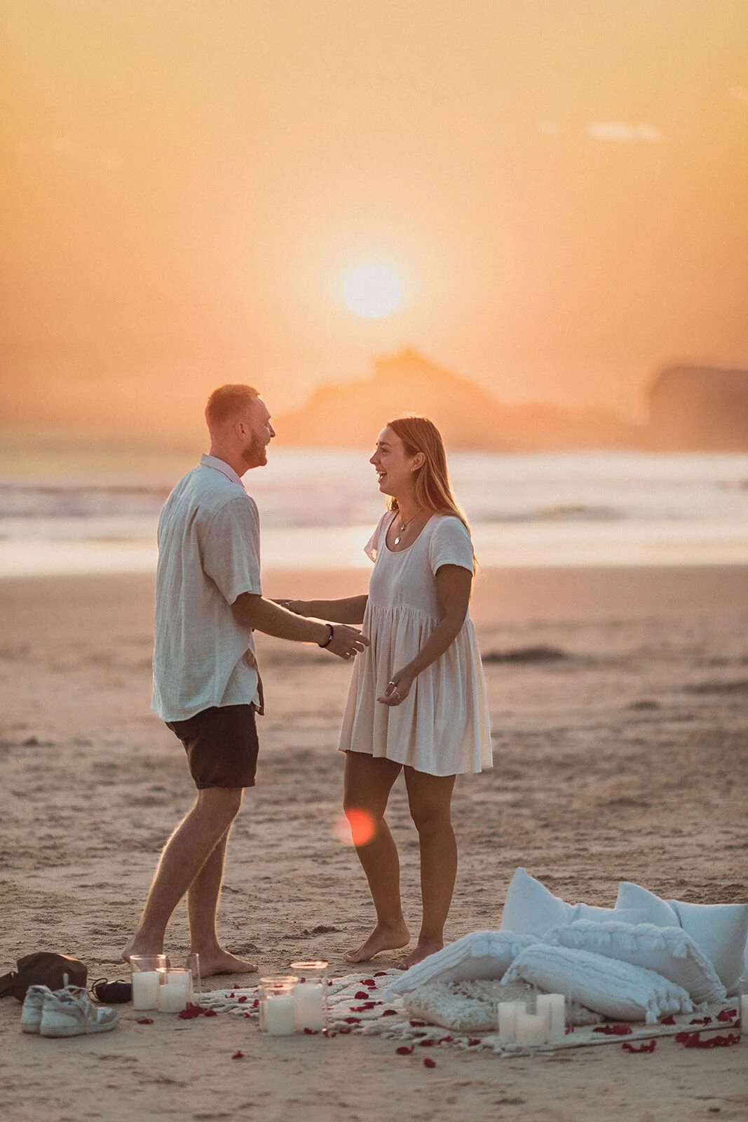 Couple facing each other and holding hands beside the pillows and candles at sunrise, Charlie and Hannah's sunrise marriage proposal at Broken Head Beach, Byron Bay
