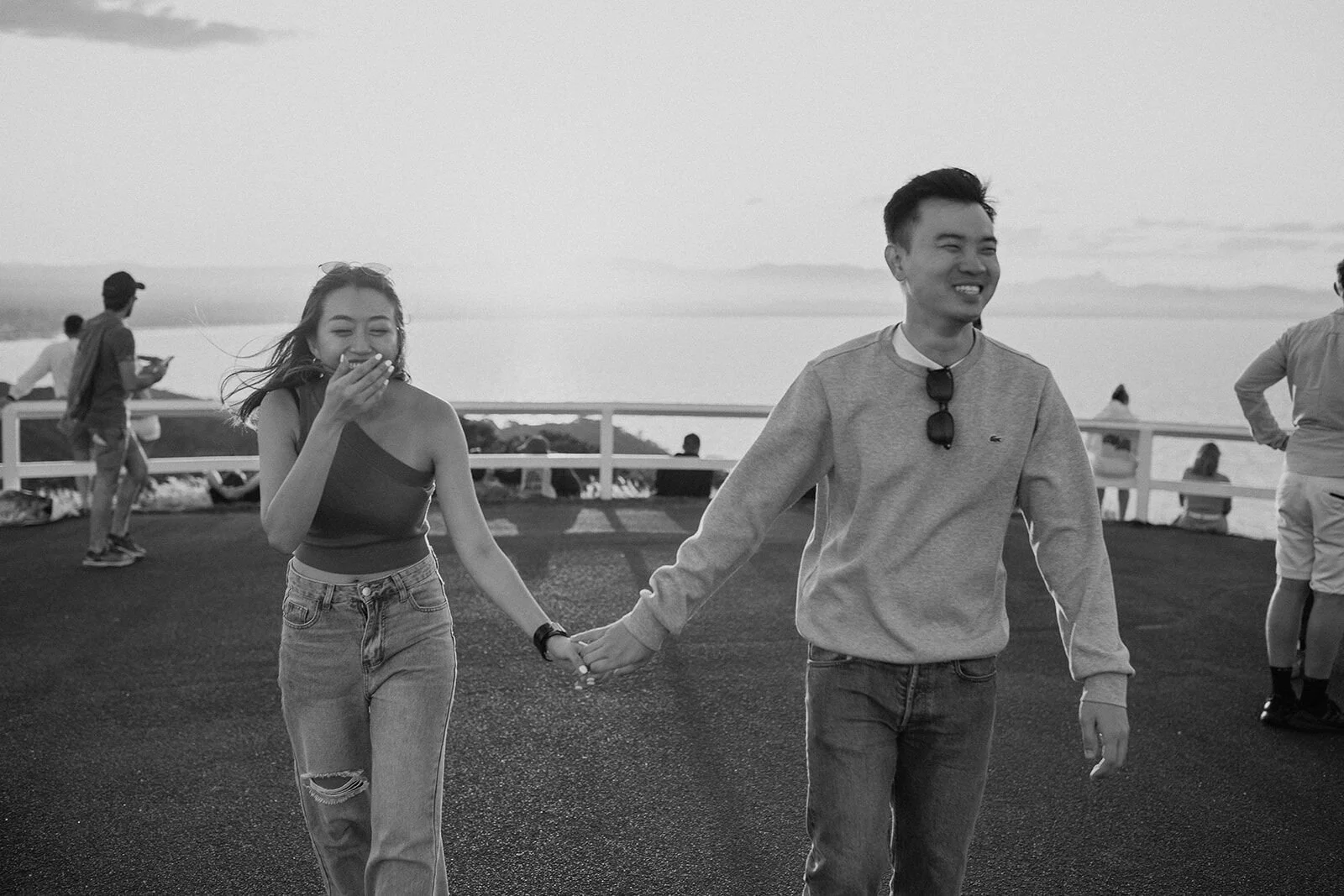 Black and white of the couple walking hand in hand and laughing, Douglas and Ying's sunset marriage proposal at Cape Byron Lighthouse, Byron Bay