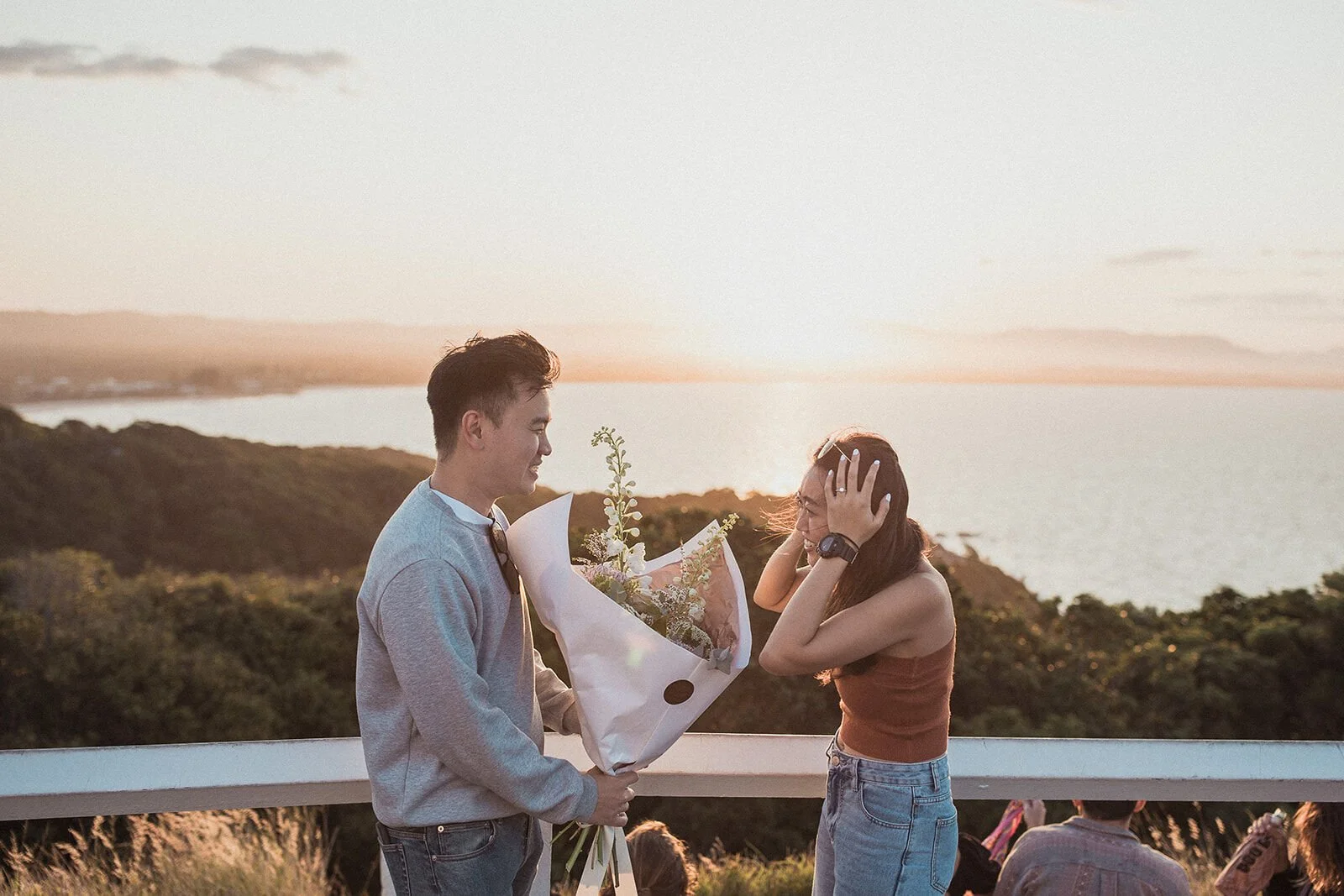Woman covering her face in delight as her partner holds a bouquet at sunset, Douglas and Ying's sunset marriage proposal at Cape Byron Lighthouse, Byron Bay