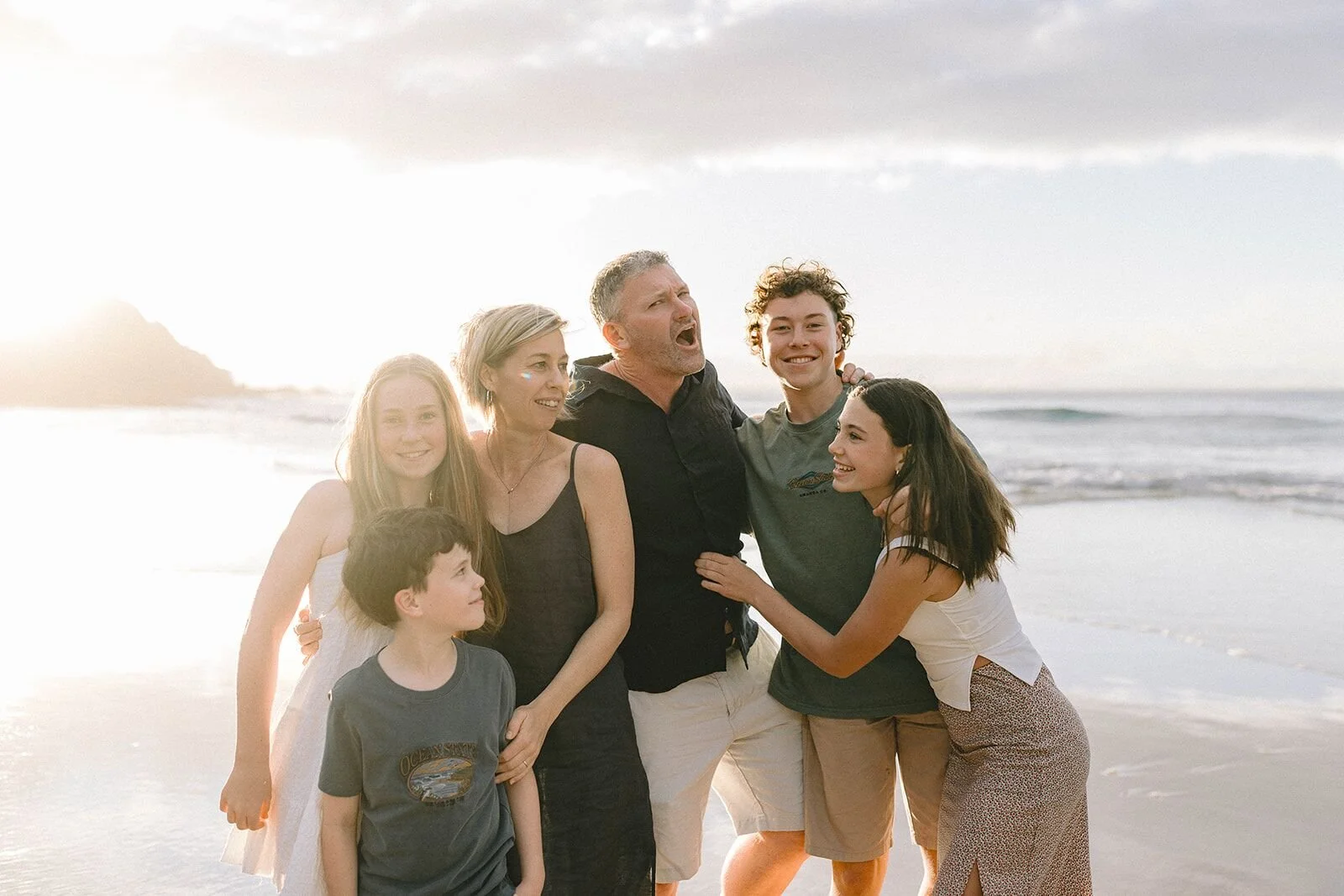 Mum, dad and their kids laughing and hugging on Wategos Beach in warm evening light