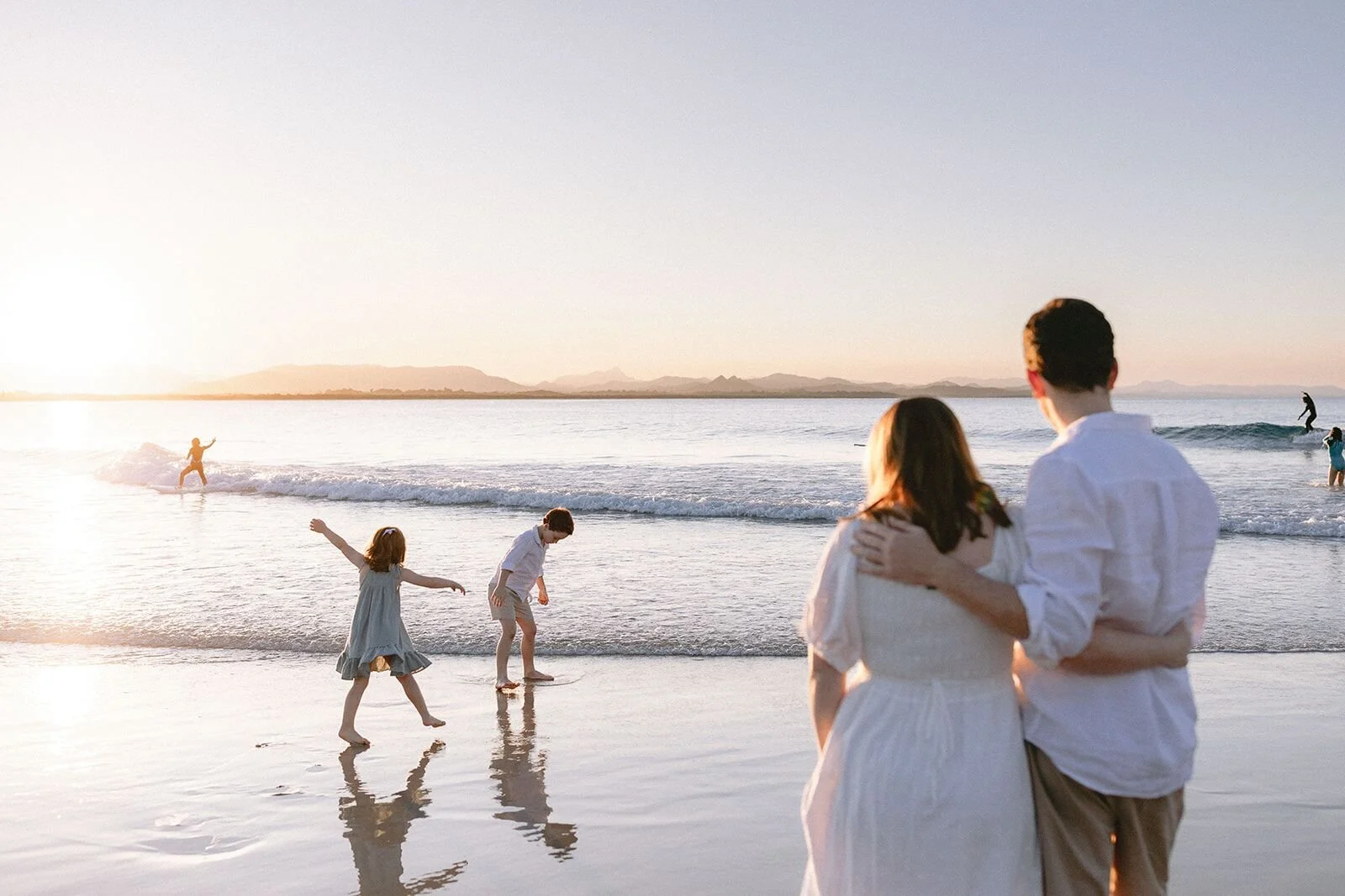 Parents with their arms around each other watching their children play in the shallows