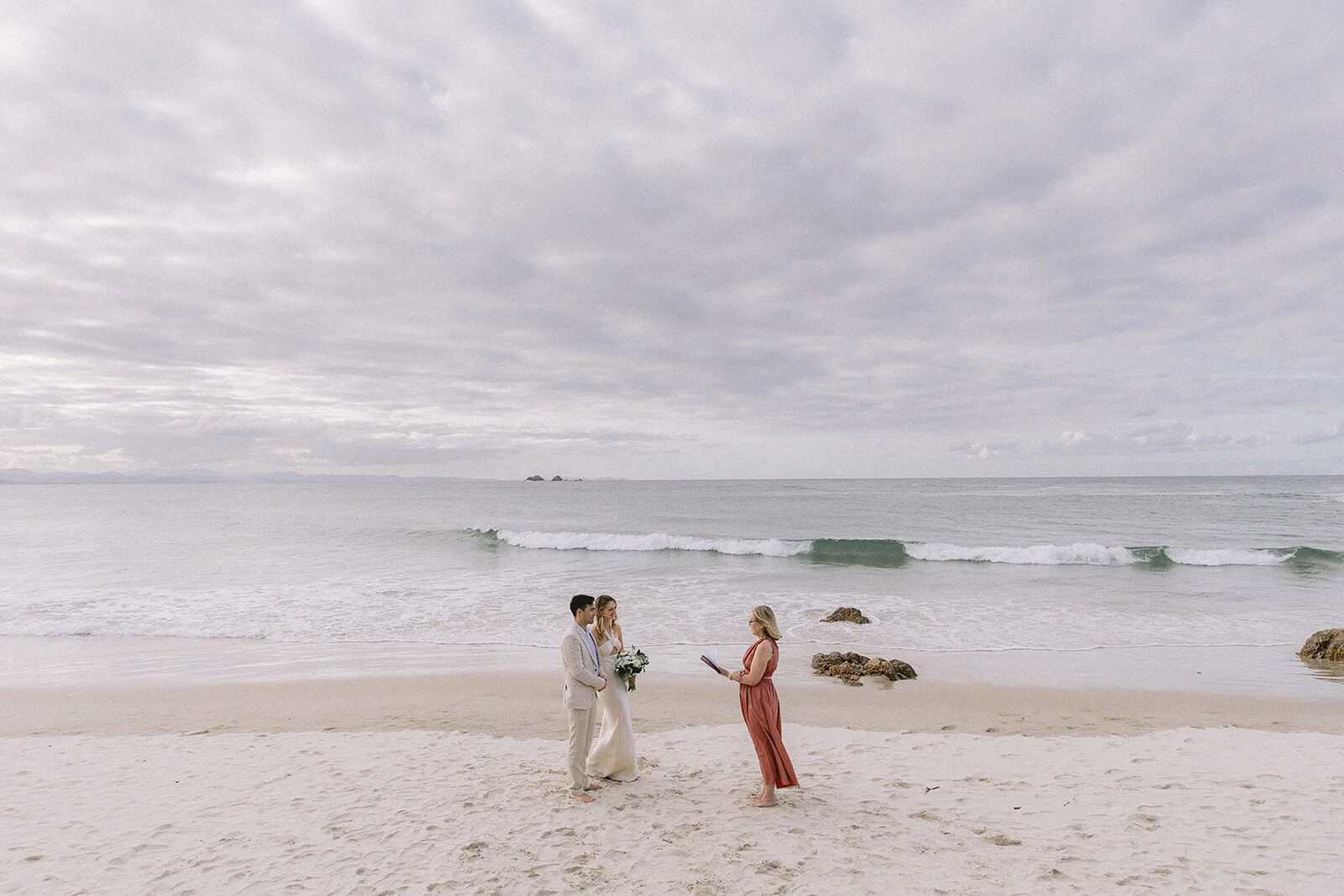 Couple and celebrant on the beach under a soft cloudy sky, Abby and James' elopement at Little Wategos Beach, Byron Bay