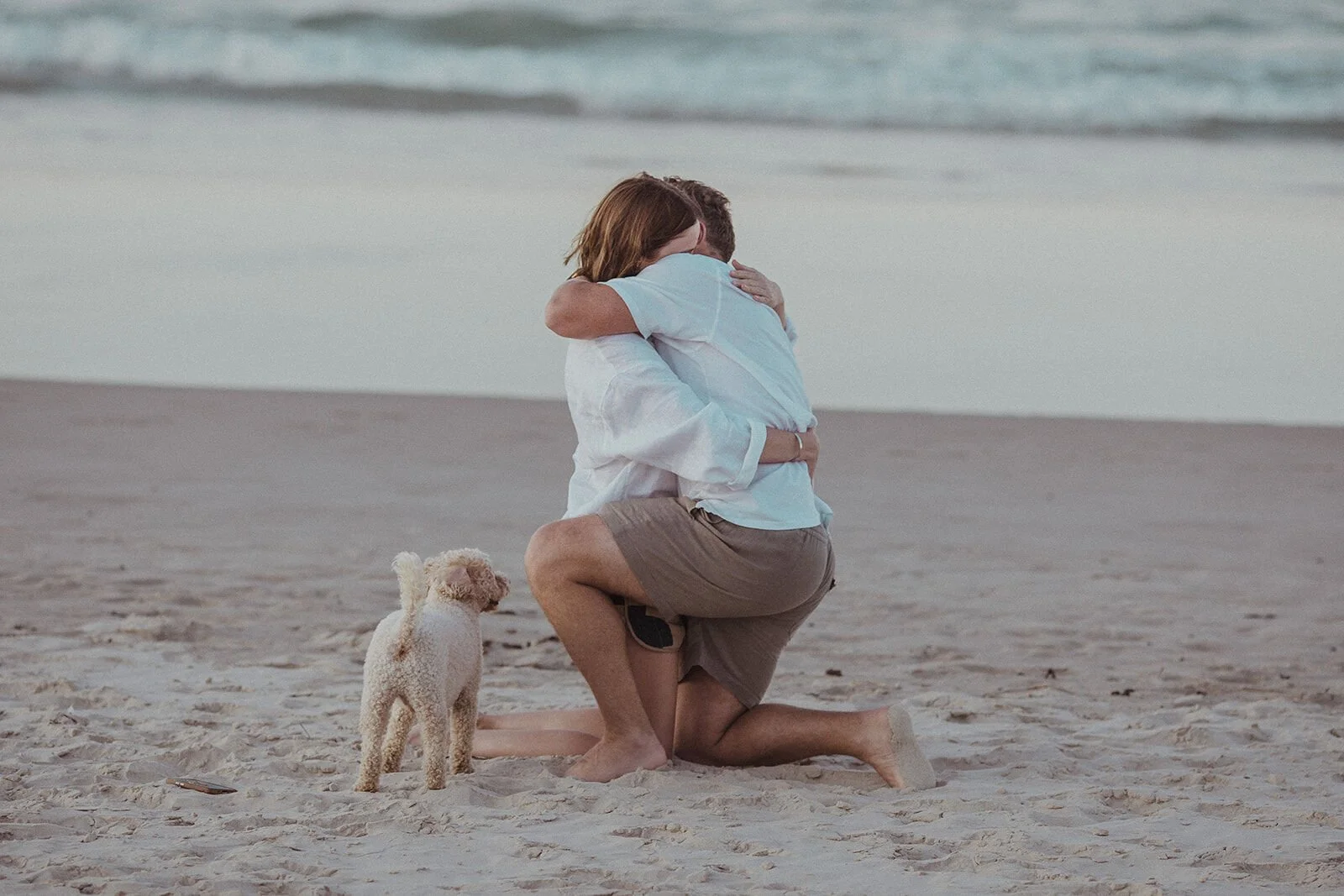Couple hugging on the beach after the proposal with their dog beside them, Jamie and Jessica's beach marriage proposal at Belongil Beach, Byron Bay