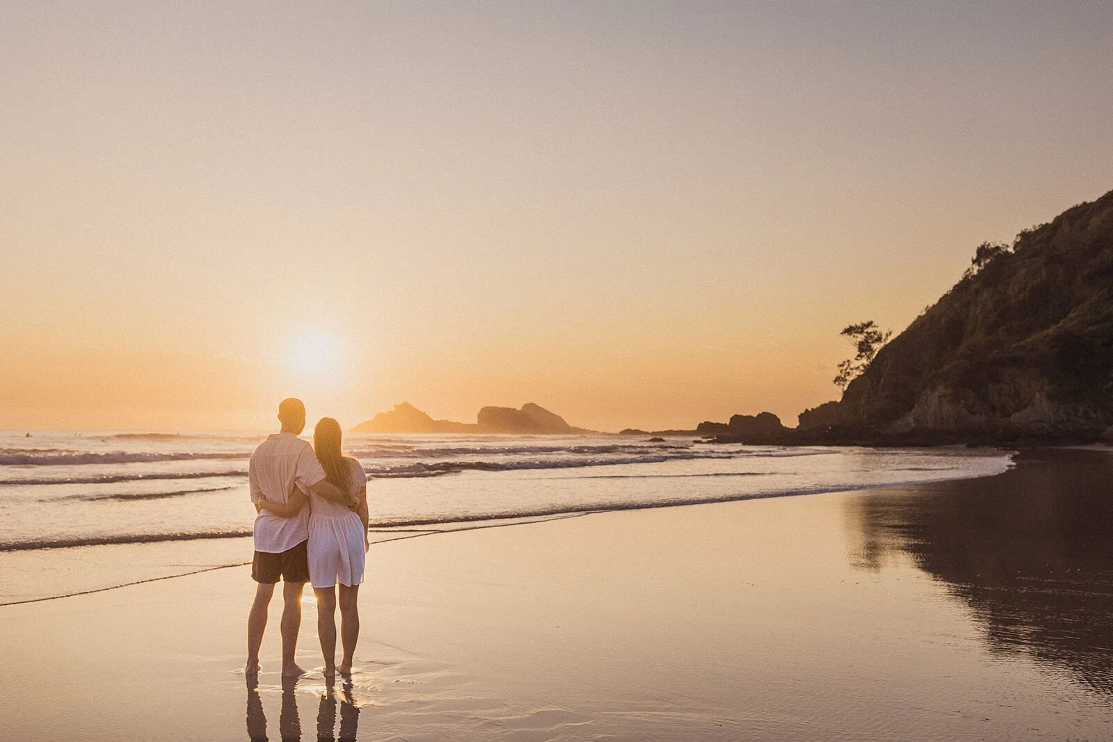Couple arm in arm from behind watching the sunrise from the shore, Charlie and Hannah's sunrise marriage proposal at Broken Head Beach, Byron Bay