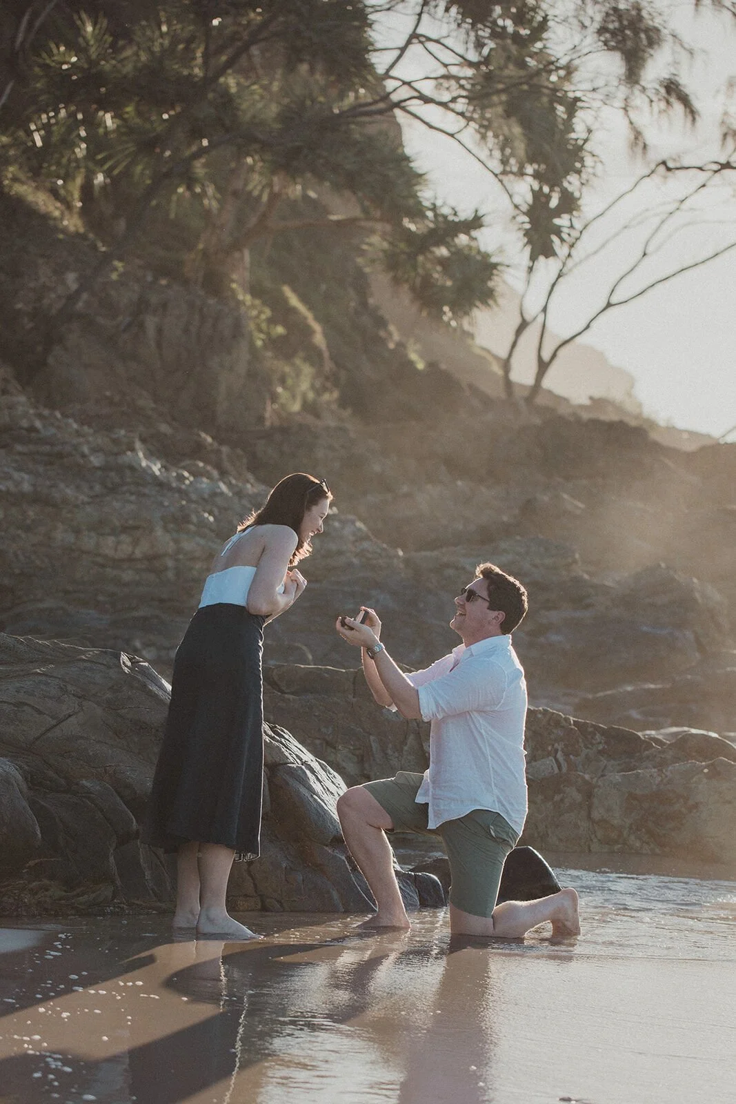 Man kneeling to propose among the rocks at the water's edge, John and Emma's sunset marriage proposal at Wategos Beach, Byron Bay