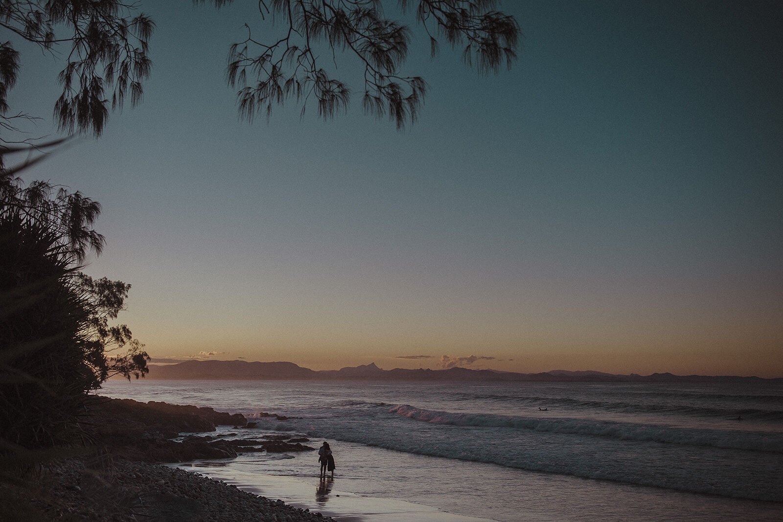Couple tiny at the shoreline at dusk with distant mountains, John and Emma's sunset marriage proposal at Wategos Beach, Byron Bay