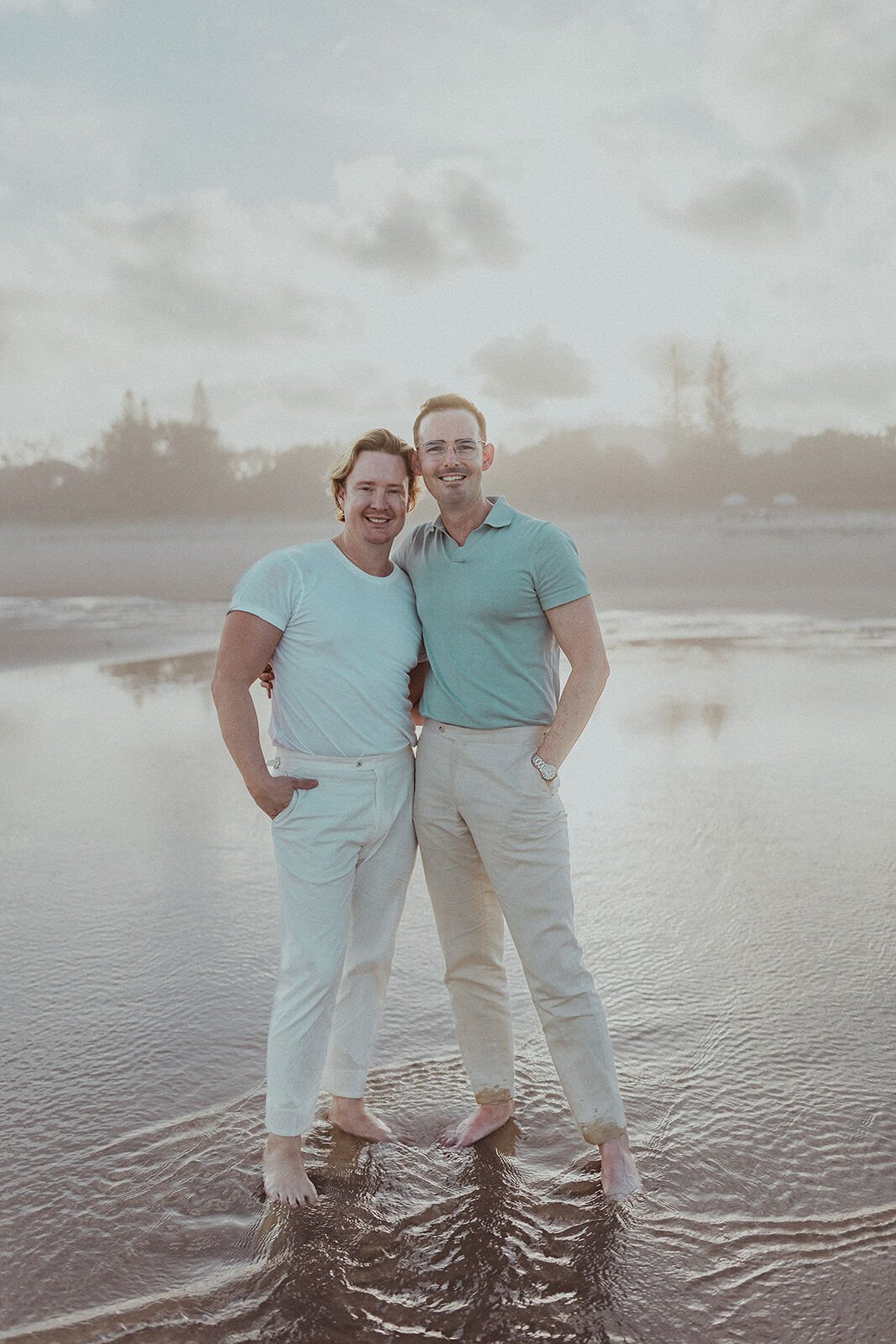 Couple standing barefoot in shallow water smiling at the camera, Byron and Luke's marriage proposal at Cabarita Beach, Northern Rivers