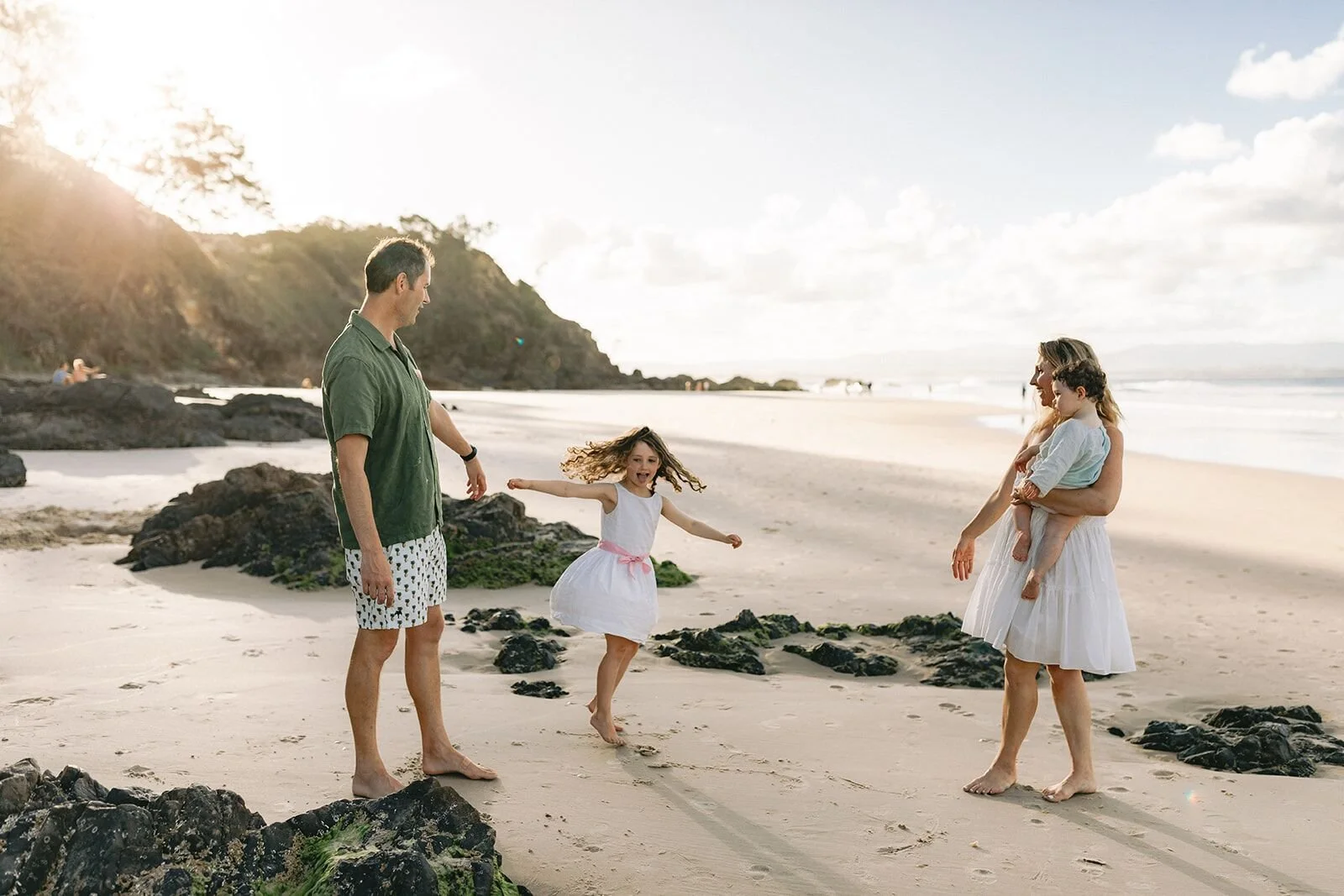 Dad and mum on the sand at Wategos with their daughter twirling between the rocks and the toddler in mum's arms