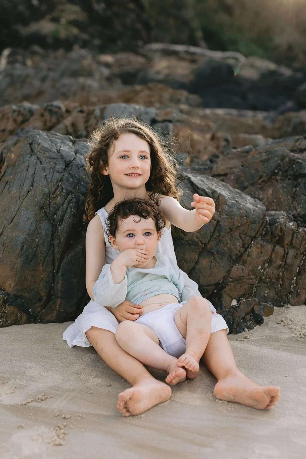 Big sister holding her baby brother on her lap, sitting against the rocks on the beach