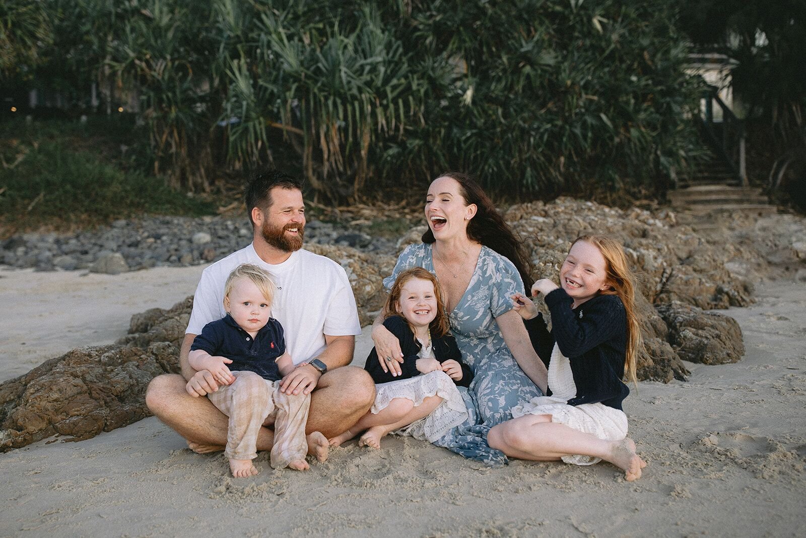 Family laughing together as they sit on the sand beside the rocks