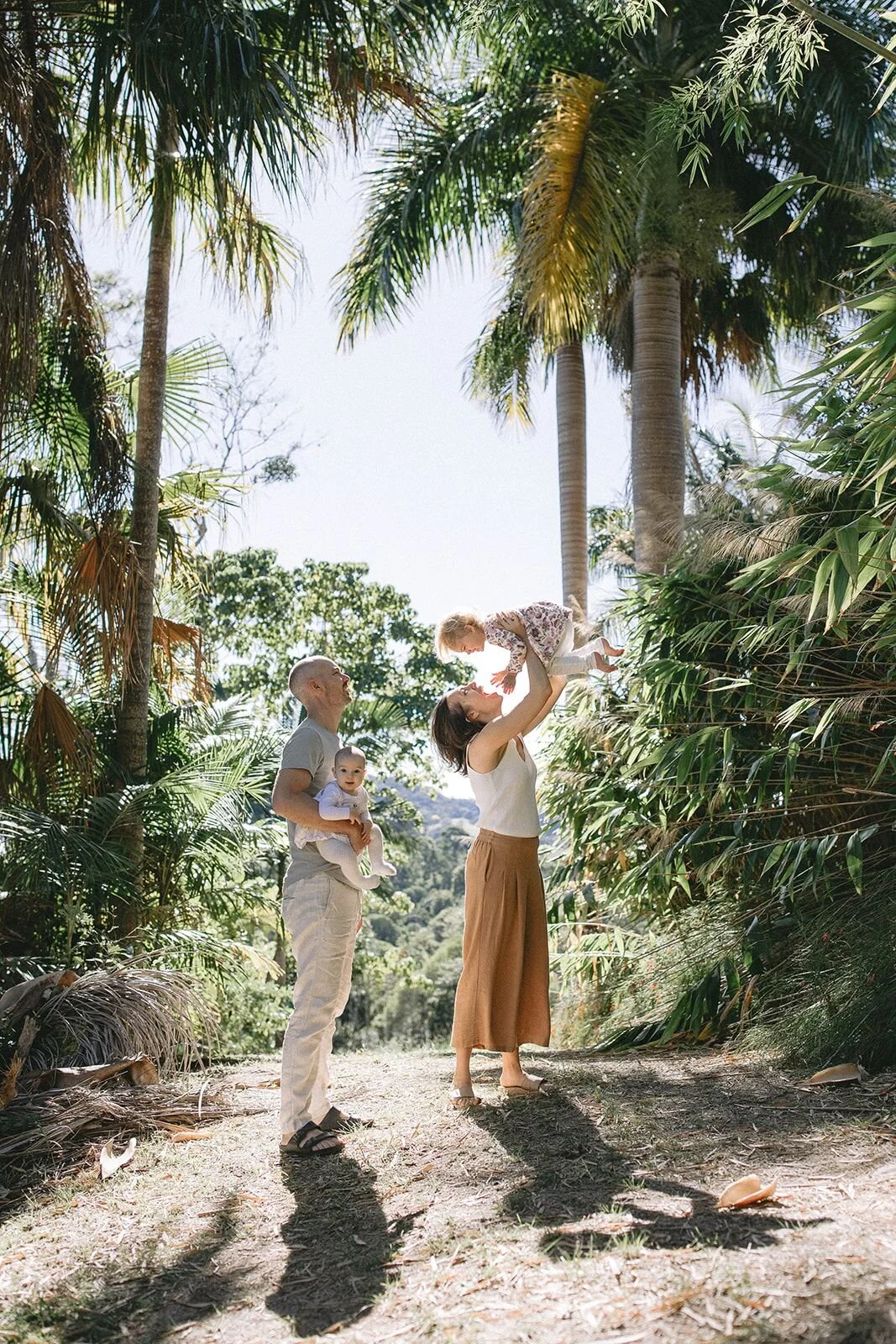 Mum lifting her baby high into the air under tall palm trees as dad looks on