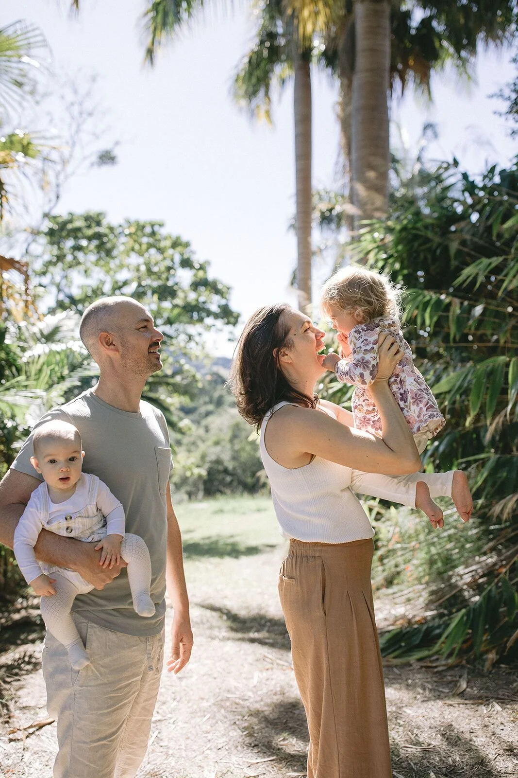 Dad holding the baby while mum lifts their toddler up for a kiss in the sunshine