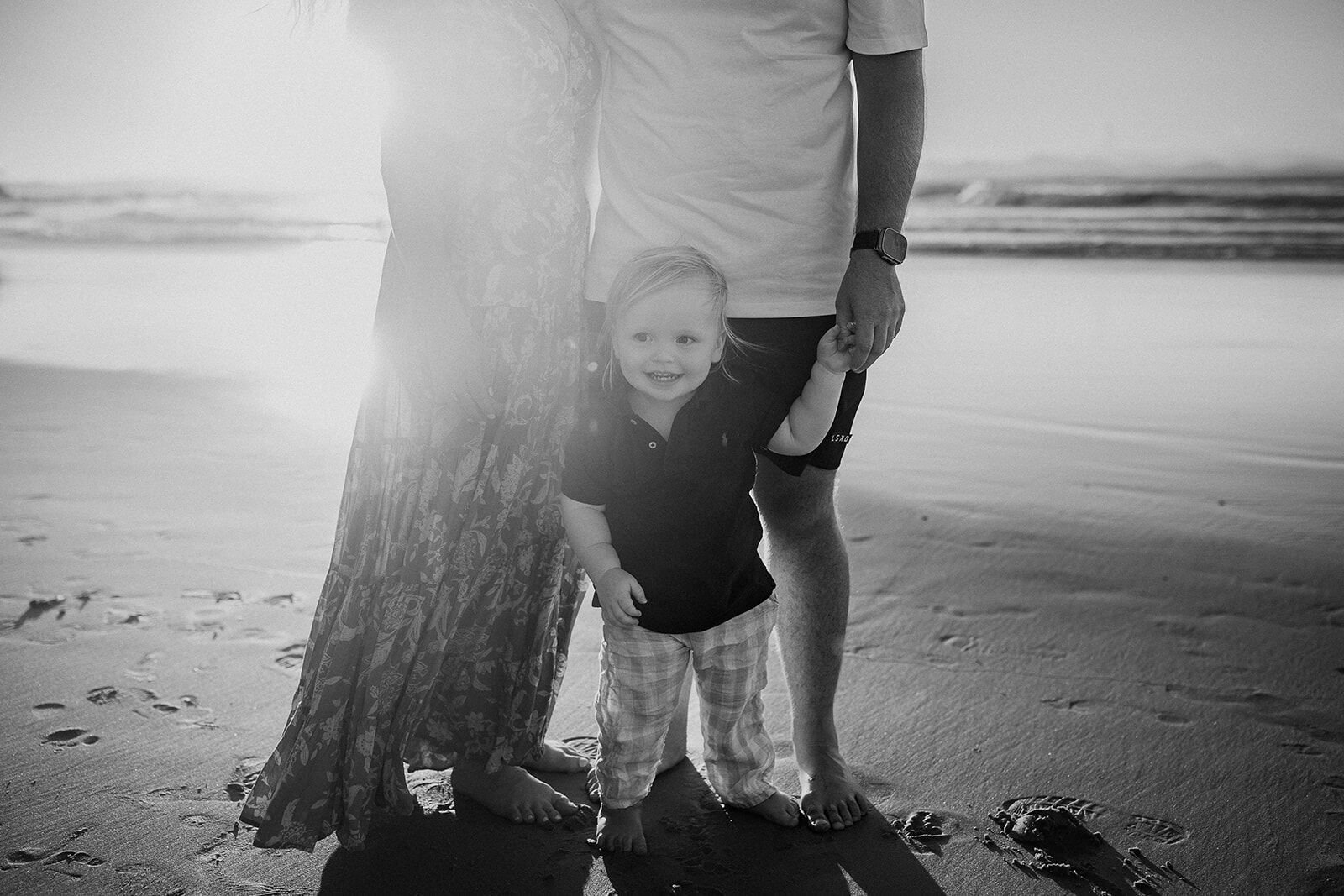 Black and white photo of a toddler holding onto his parents' legs on the beach