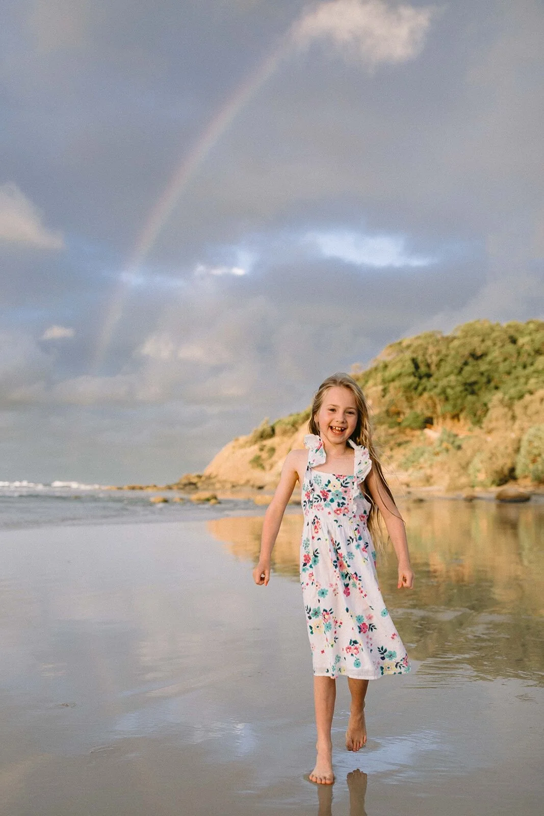 Girl in a floral dress walking across the wet sand with a rainbow behind her, Wategos Beach