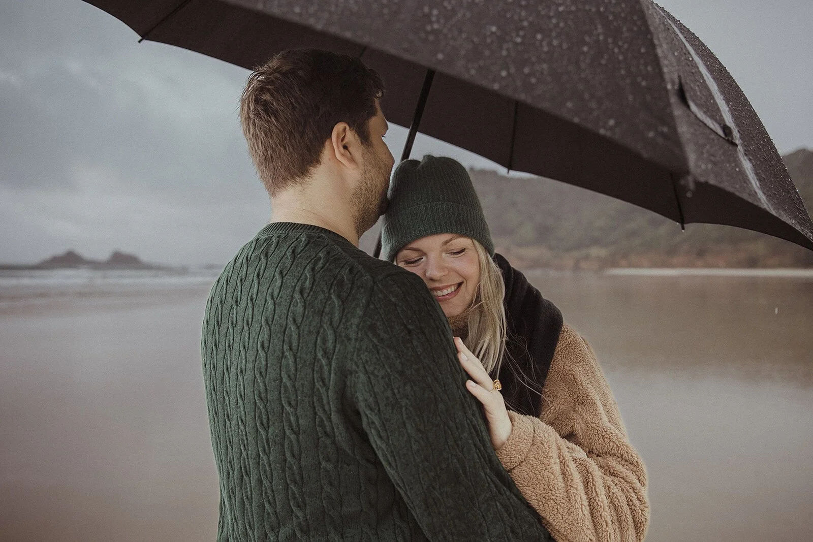 Couple holding each other close under the umbrella in the rain, Raphael and Vany's rainy day marriage proposal at Broken Head Beach, Byron Bay