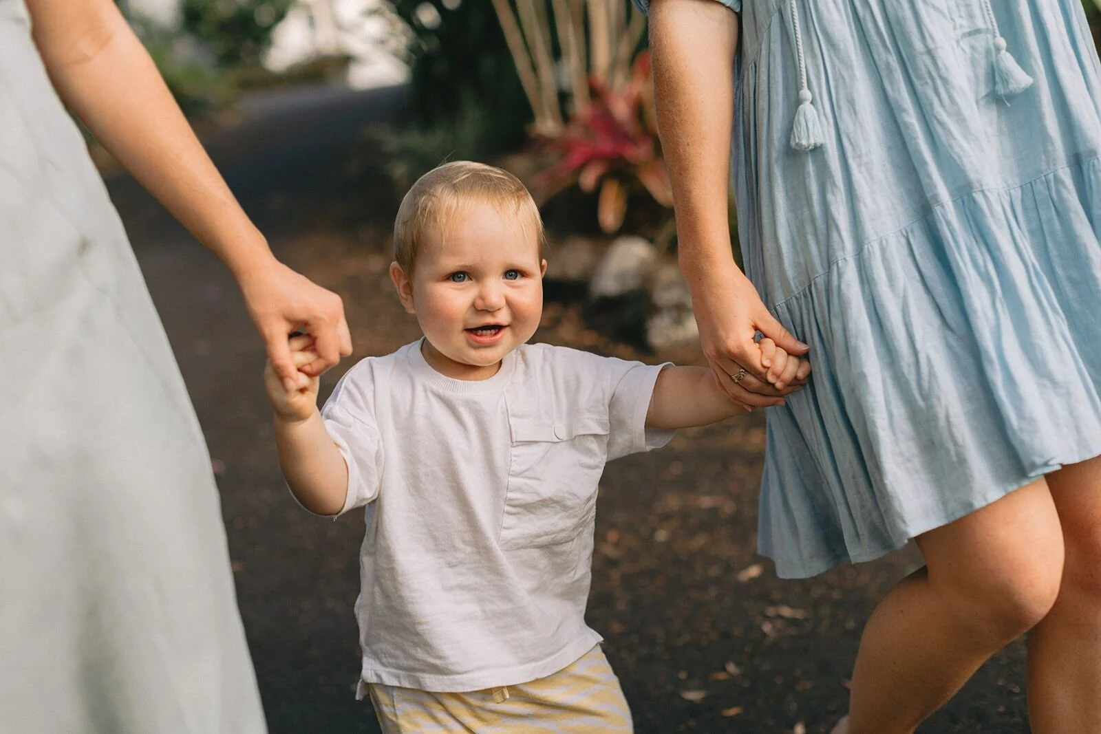 Smiling toddler in a white t-shirt holding hands with two family members as they walk