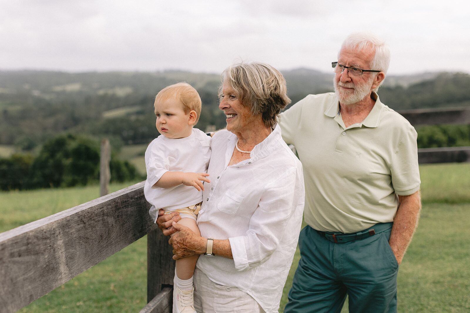 Grandparents smiling with their baby grandson on a hillside farm at Christmas