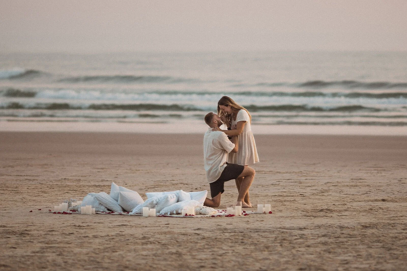 Woman leaning in to hold her partner's face as he kneels on the sand, Charlie and Hannah's sunrise marriage proposal at Broken Head Beach, Byron Bay