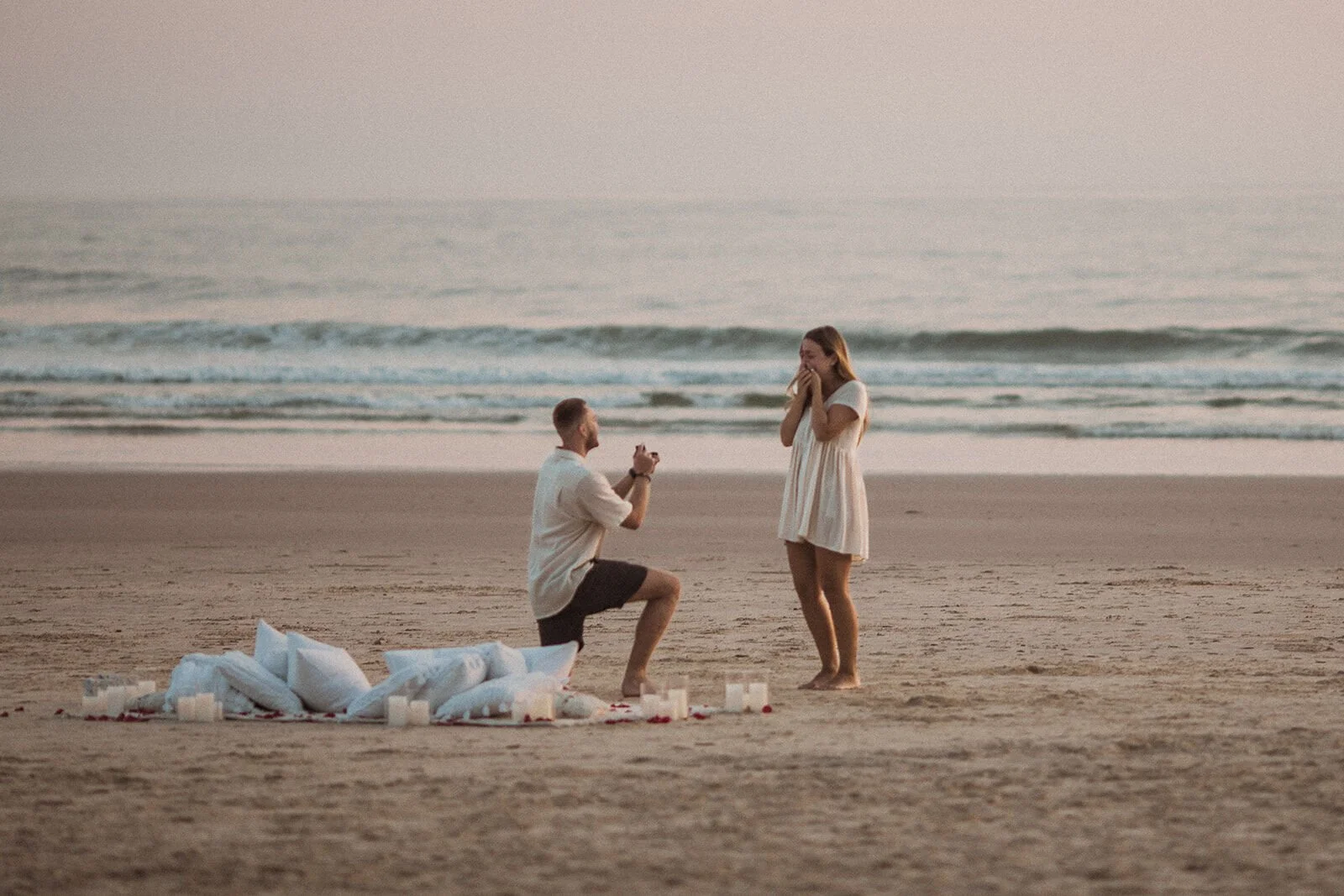Man on one knee proposing on the beach as the woman covers her mouth, with pillows and candles behind them, Charlie and Hannah's sunrise marriage proposal at Broken Head Beach, Byron Bay