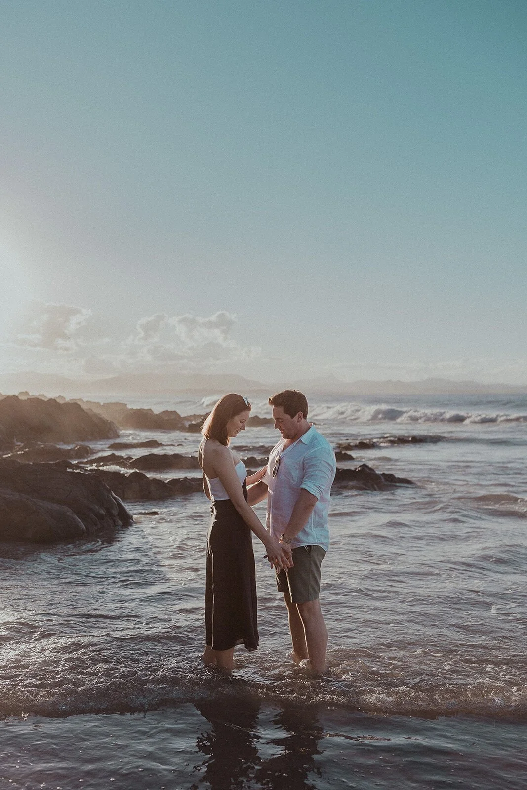 Couple standing hand in hand in the shallow water, John and Emma's sunset marriage proposal at Wategos Beach, Byron Bay
