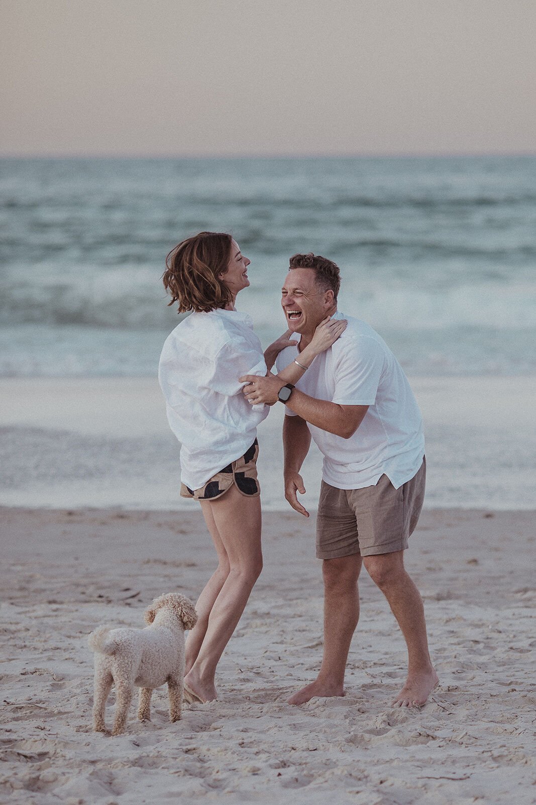 Couple laughing together as their dog jumps up at them, Jamie and Jessica's beach marriage proposal at Belongil Beach, Byron Bay