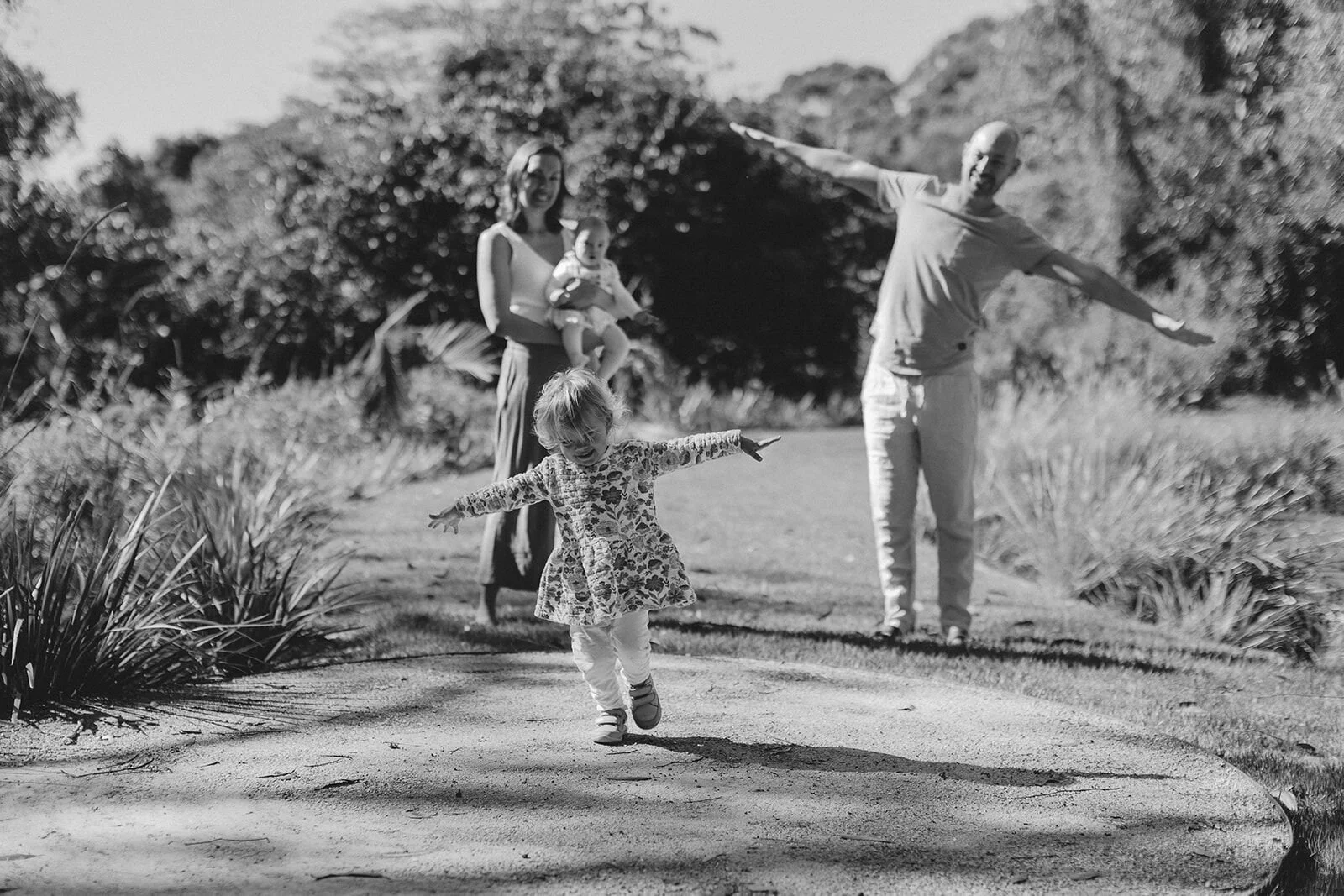 Black and white photo of a toddler dancing on a garden path with her arms out, parents behind her