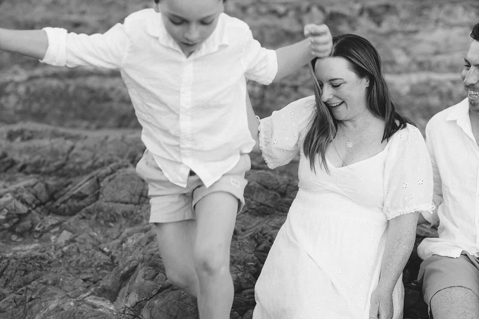 Black and white photo of a boy climbing over the rocks beside his smiling mum