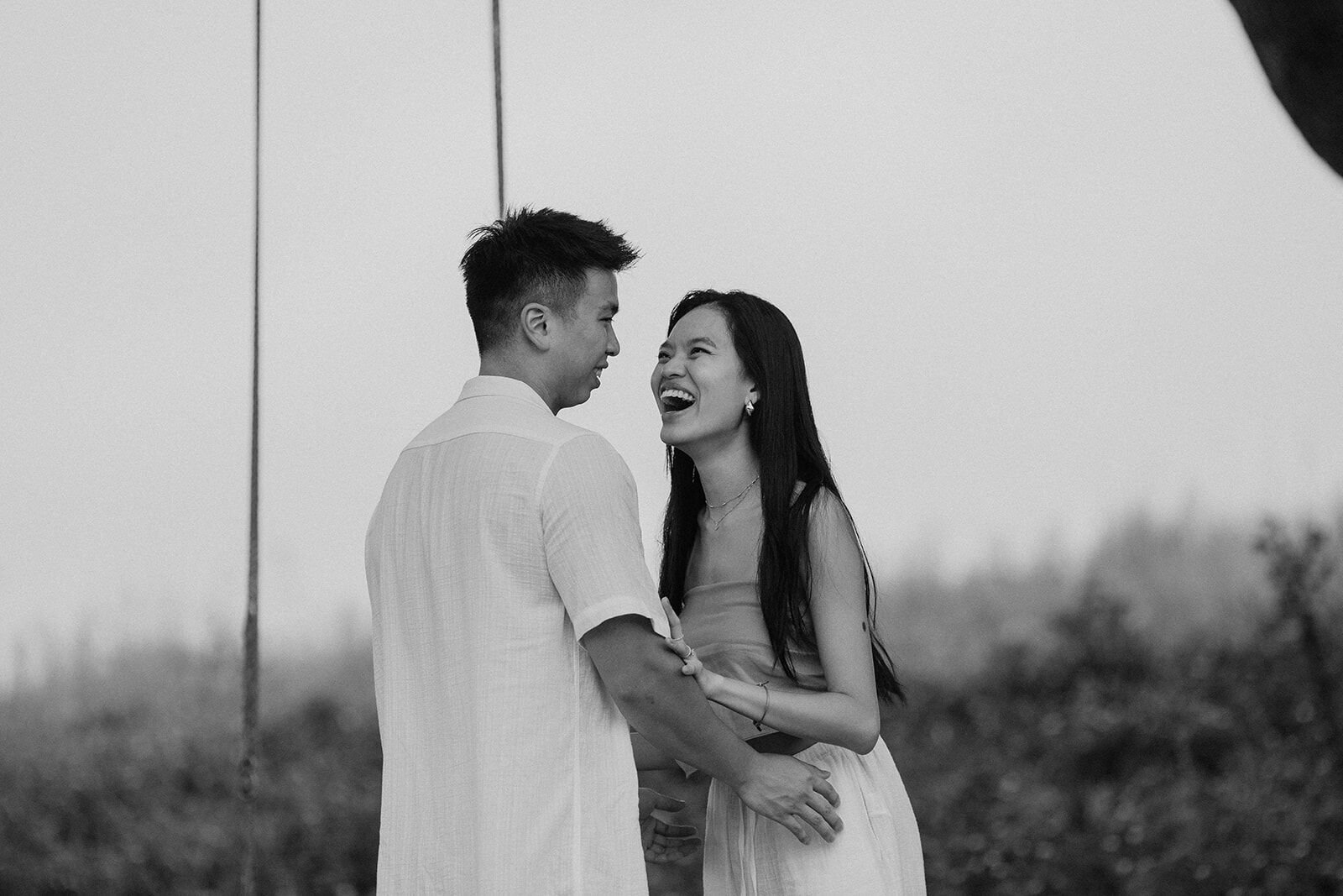 Black and white of the couple laughing together beside the rope swing, Darien and Jacqui's picnic marriage proposal at Heartwood Farm, Byron Bay hinterland
