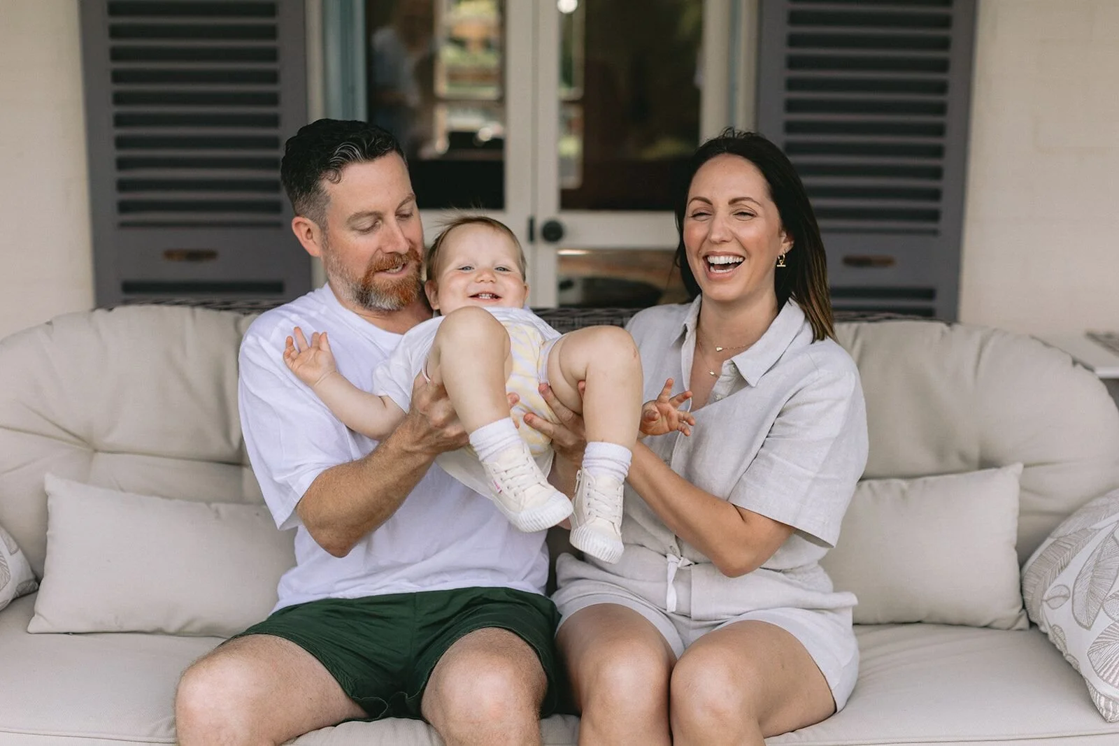 Dad tickling his giggling toddler on his lap while mum laughs beside them