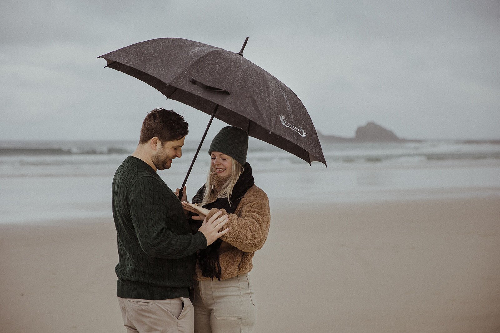 Couple admiring the ring under a black umbrella with a rock stack in the sea behind, Raphael and Vany's rainy day marriage proposal at Broken Head Beach, Byron Bay