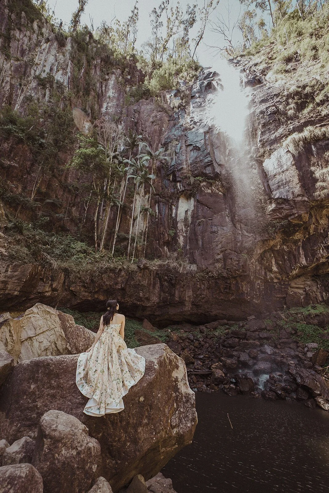 Bride in a floral dress on a rock beside a waterfall, Maggie and William's pre-wedding photos in Byron Bay